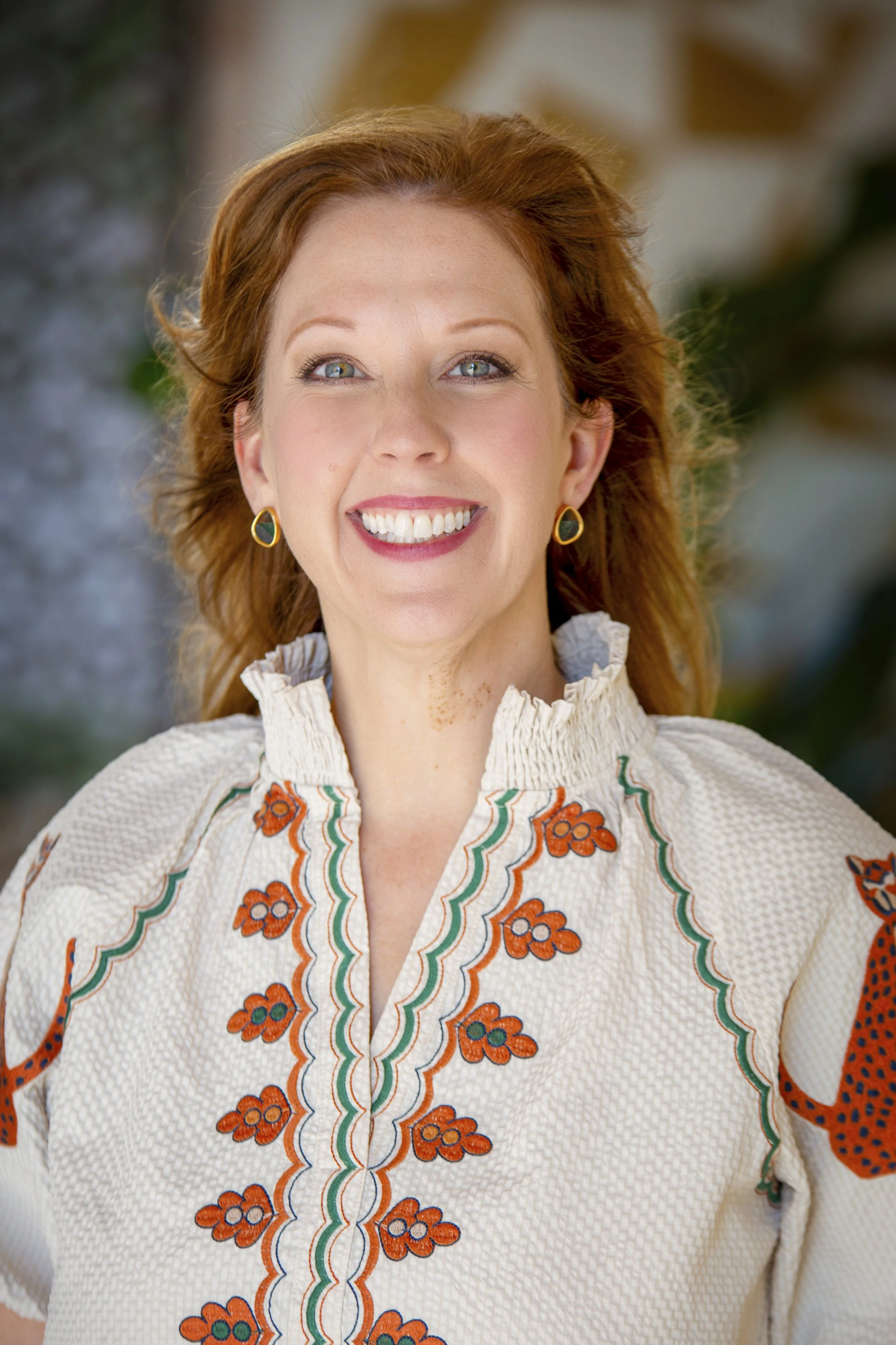 Portrait of a smiling woman with red hair, wearing earrings and a cream-colored embroidered blouse, standing outdoors with blurred background.