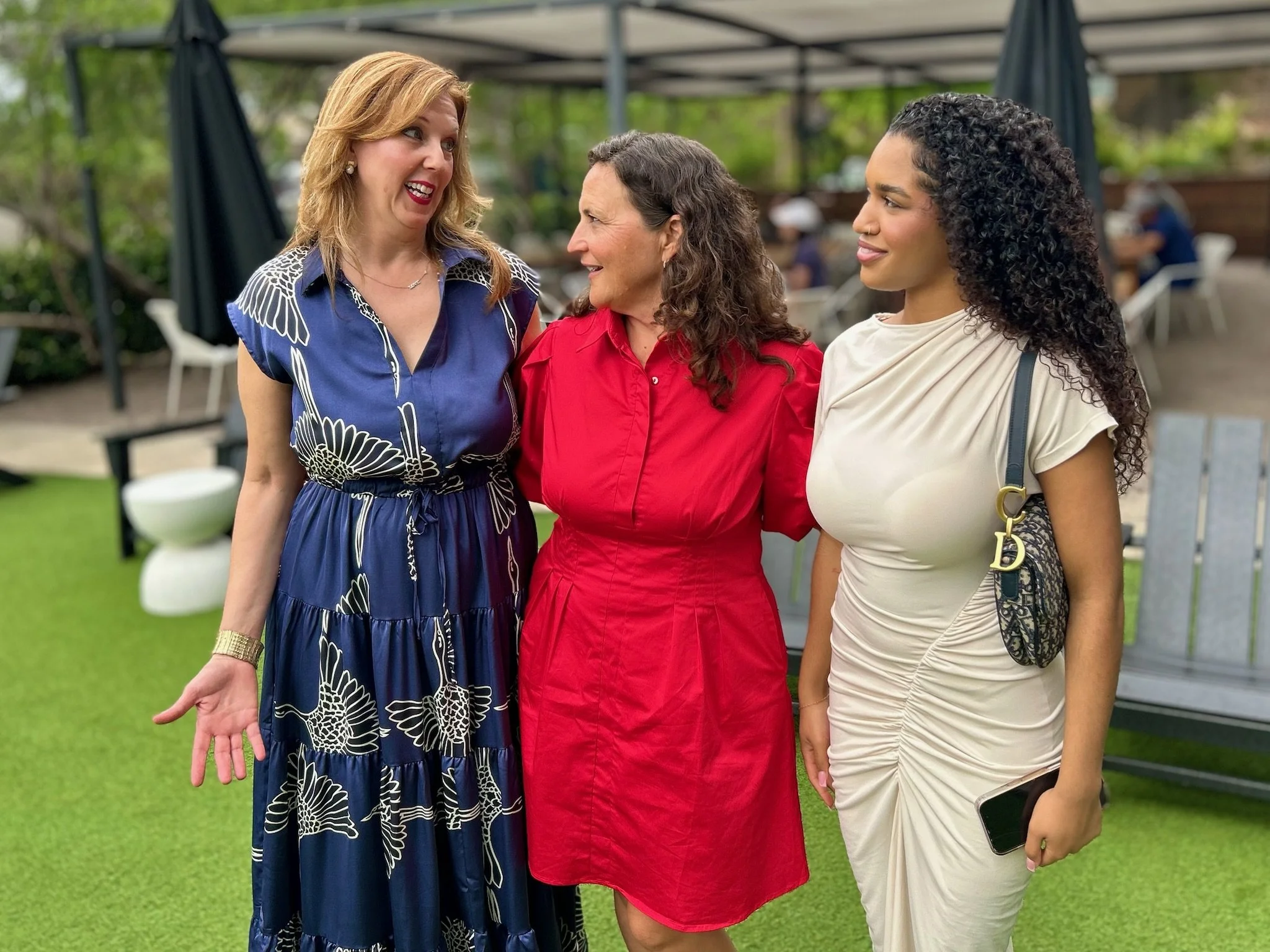 Three women standing close together outdoors, smiling and talking. One woman is wearing a blue dress with white patterns, another in a red dress, and the third in a cream-colored dress holding a smartphone. They are under a canopy with a green background and other people sitting at tables in the distance.