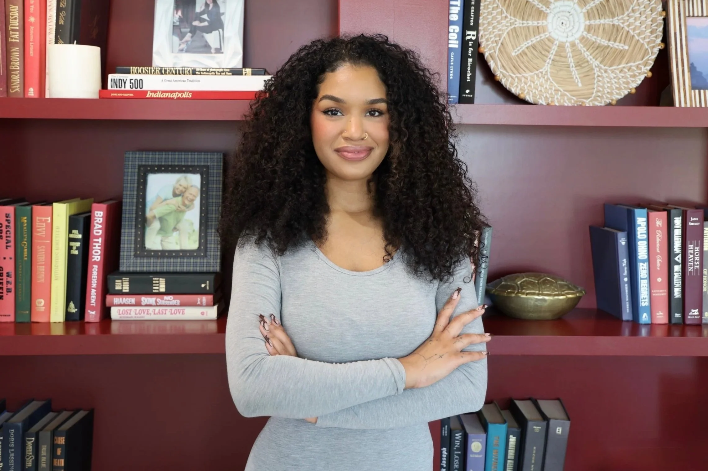 A woman with curly hair standing in a modern, well-lit living room, wearing a black suit with a white top and white heels.