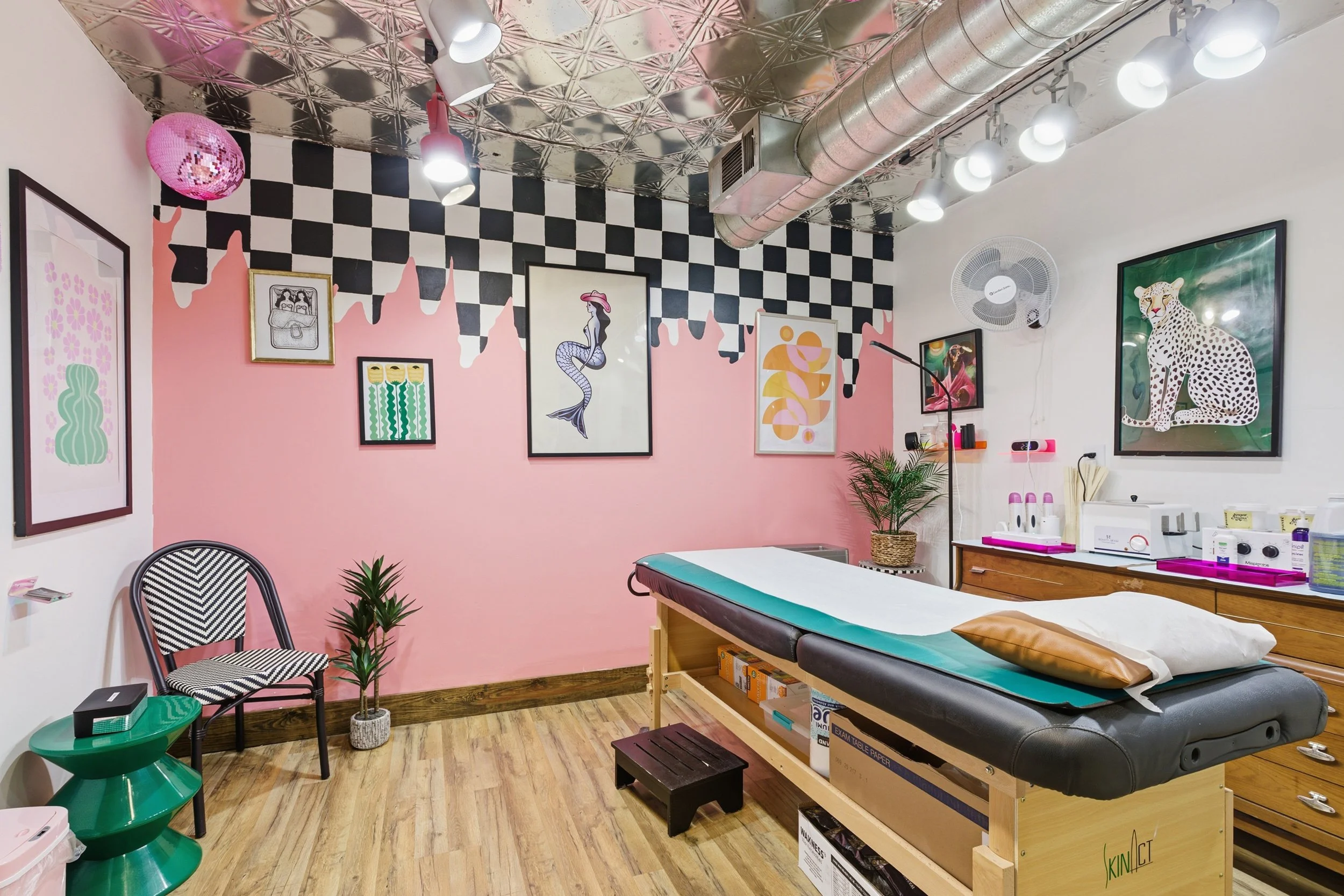 A room with pink and black checkered wall art, framed pictures of abstract and animal designs, and a pink paper lantern. There is a massage or examination table with a pillow, a wooden storage unit, and a small black stool. The ceiling has metallic t