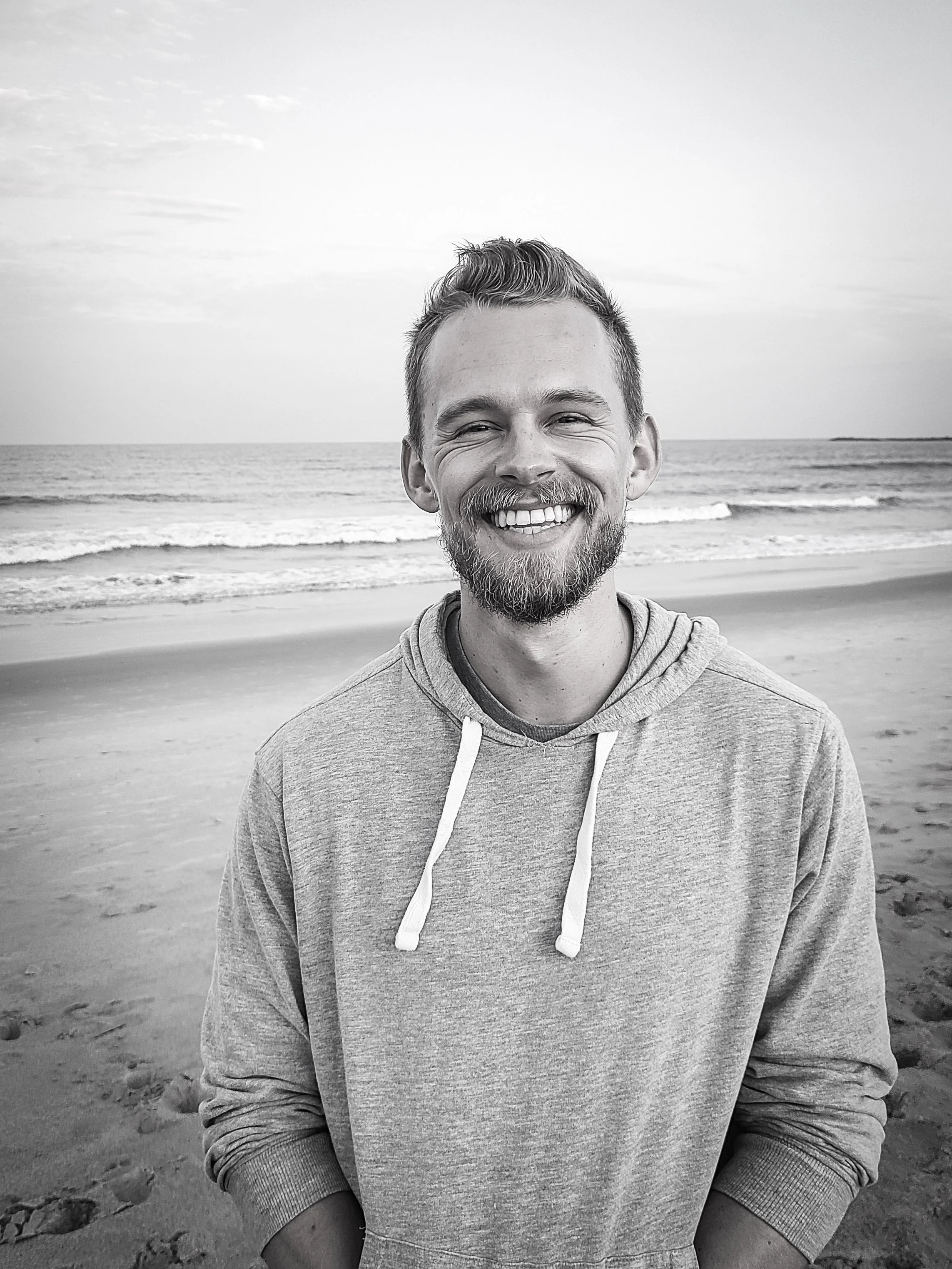 A smiling man with a beard and mustache wearing a hoodie, standing on a beach with the ocean and sky in the background.