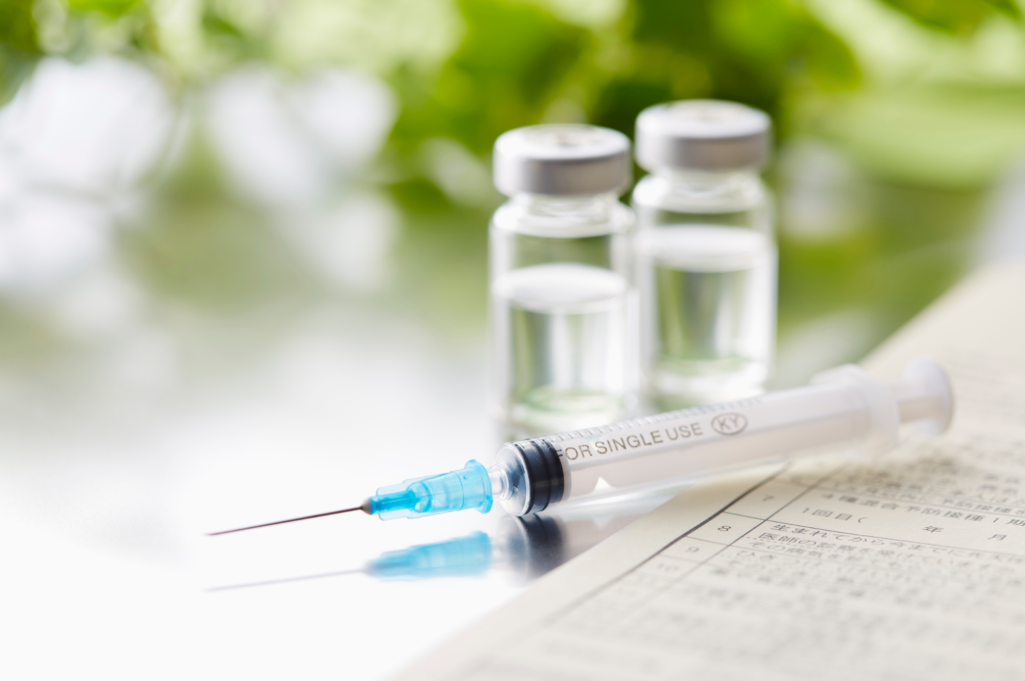 B12 & Homeopathic Injection vials and a syringe on a table with paper and blurred green background.