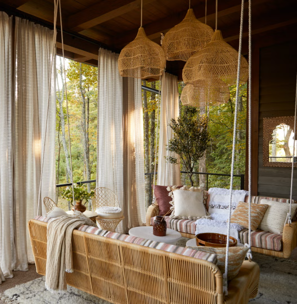 Sunlit indoor living room with rattan hanging lamps, wicker furniture, striped cushions, potted plants, and view of trees outside through large windows with white curtains.