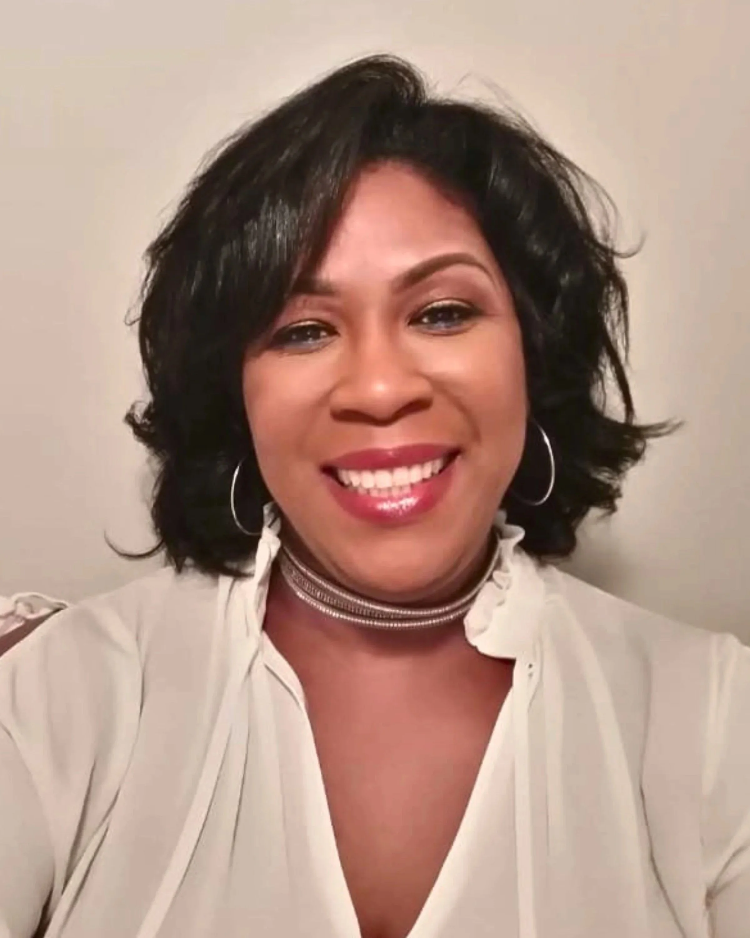 Close-up of a smiling woman with short black hair, wearing a cream-colored blouse and hoop earrings, standing against a plain wall.