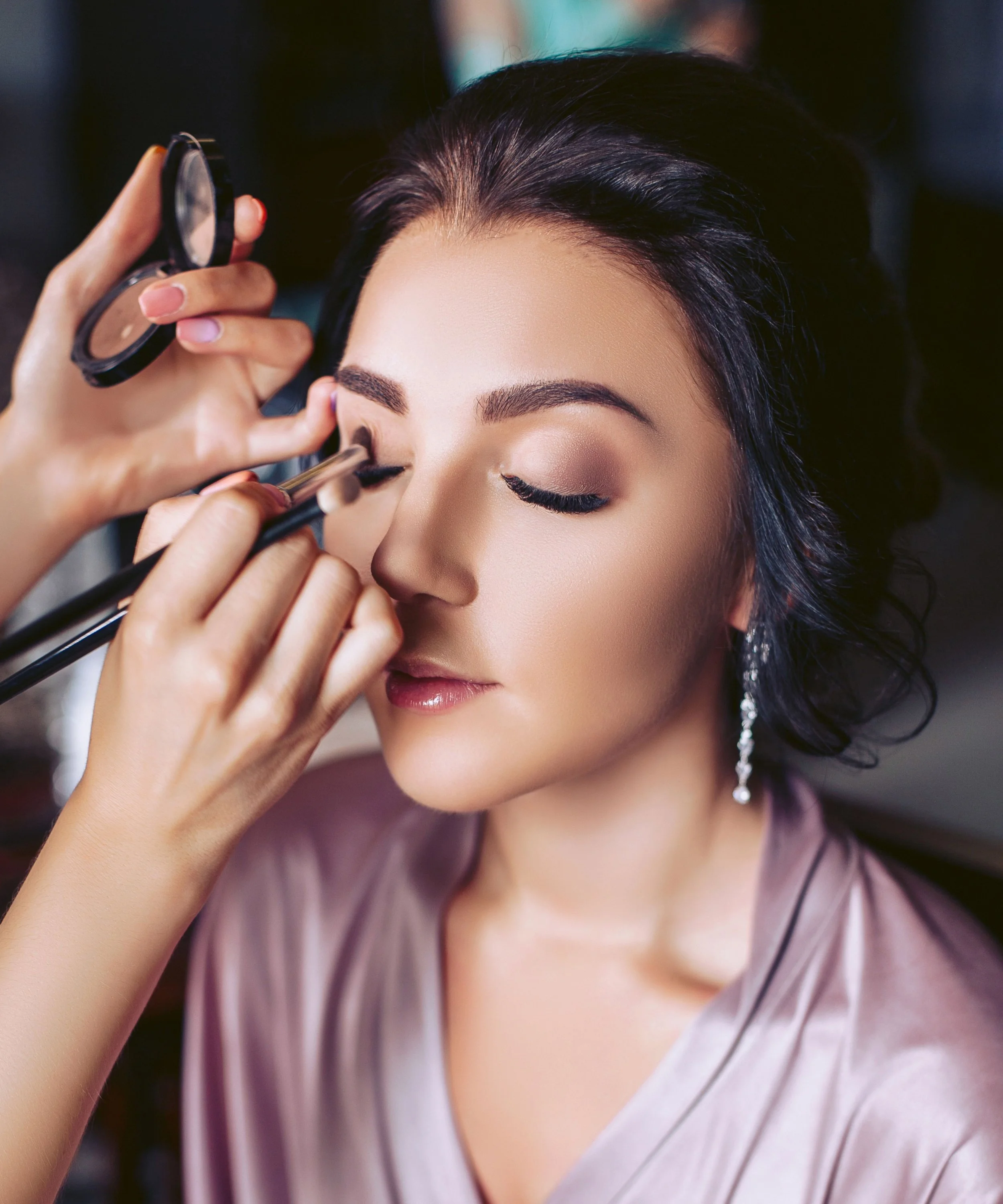 A woman with closed eyes having her makeup done by a makeup artist.