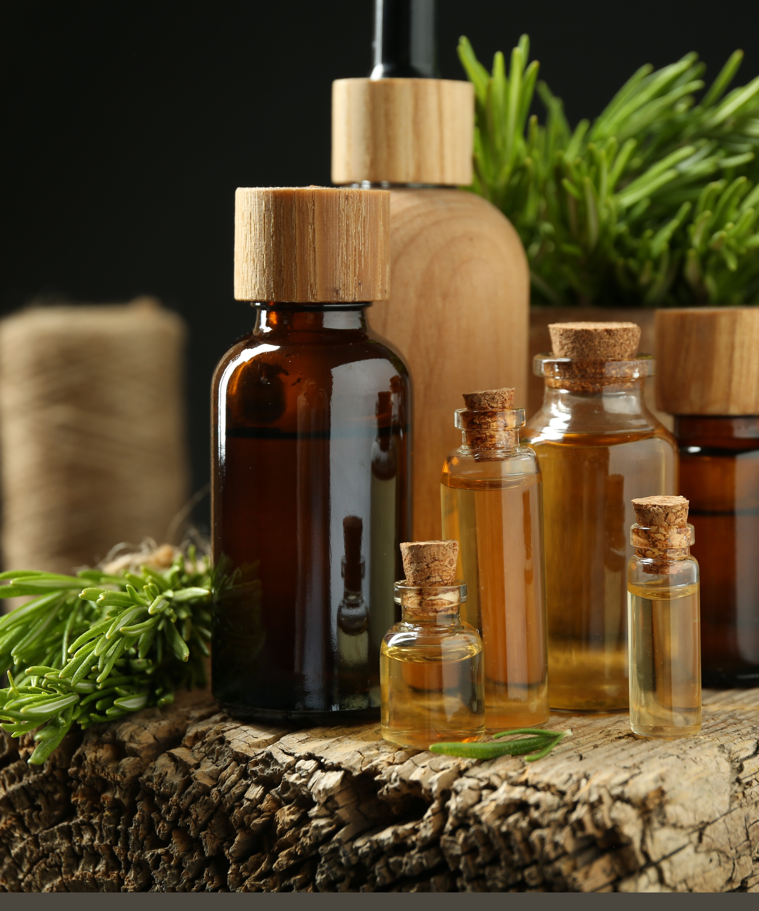 Assorted small glass and amber bottles with cork stoppers containing oils, arranged on a rustic wooden surface with green herbs and wooden spoons in the background.