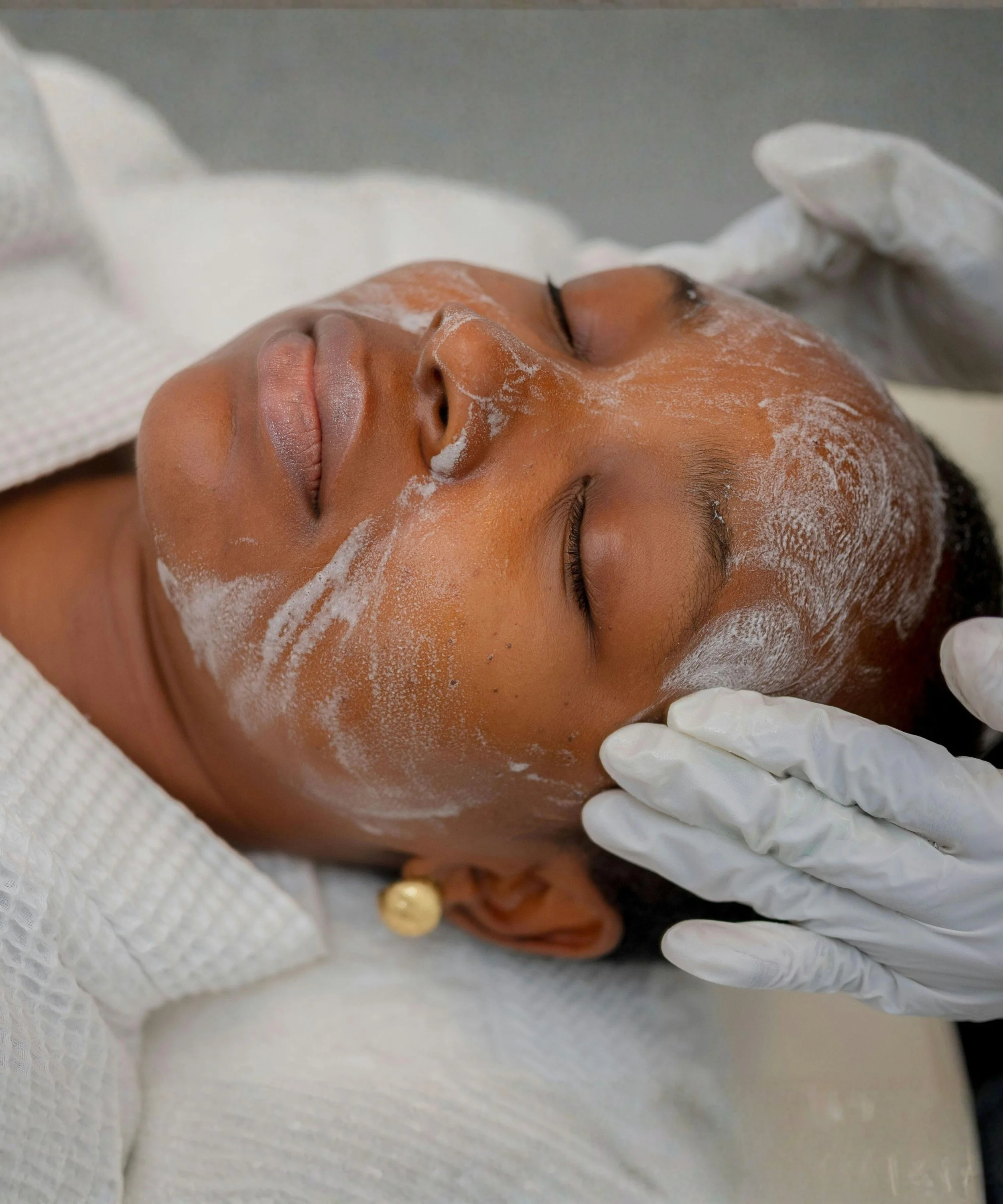 Woman receiving a facial treatment with a foamy cleanser on her face, lying down with eyes closed, wearing a white robe and earrings.