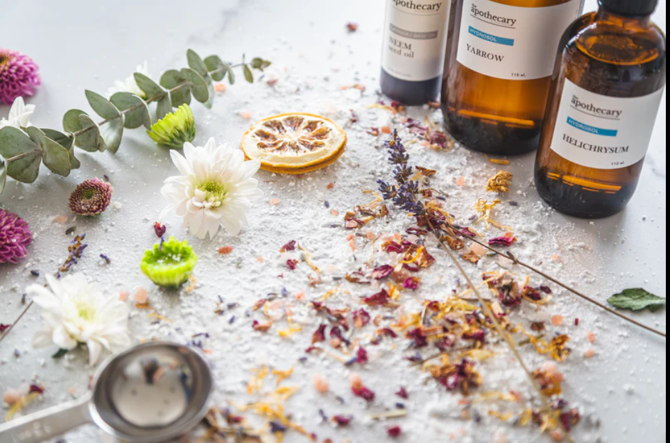 A flat lay of herbal and aromatherapy supplies including dried flower petals, essential oil bottles, flowers, dried citrus slice, and a metal scoop on a white surface.