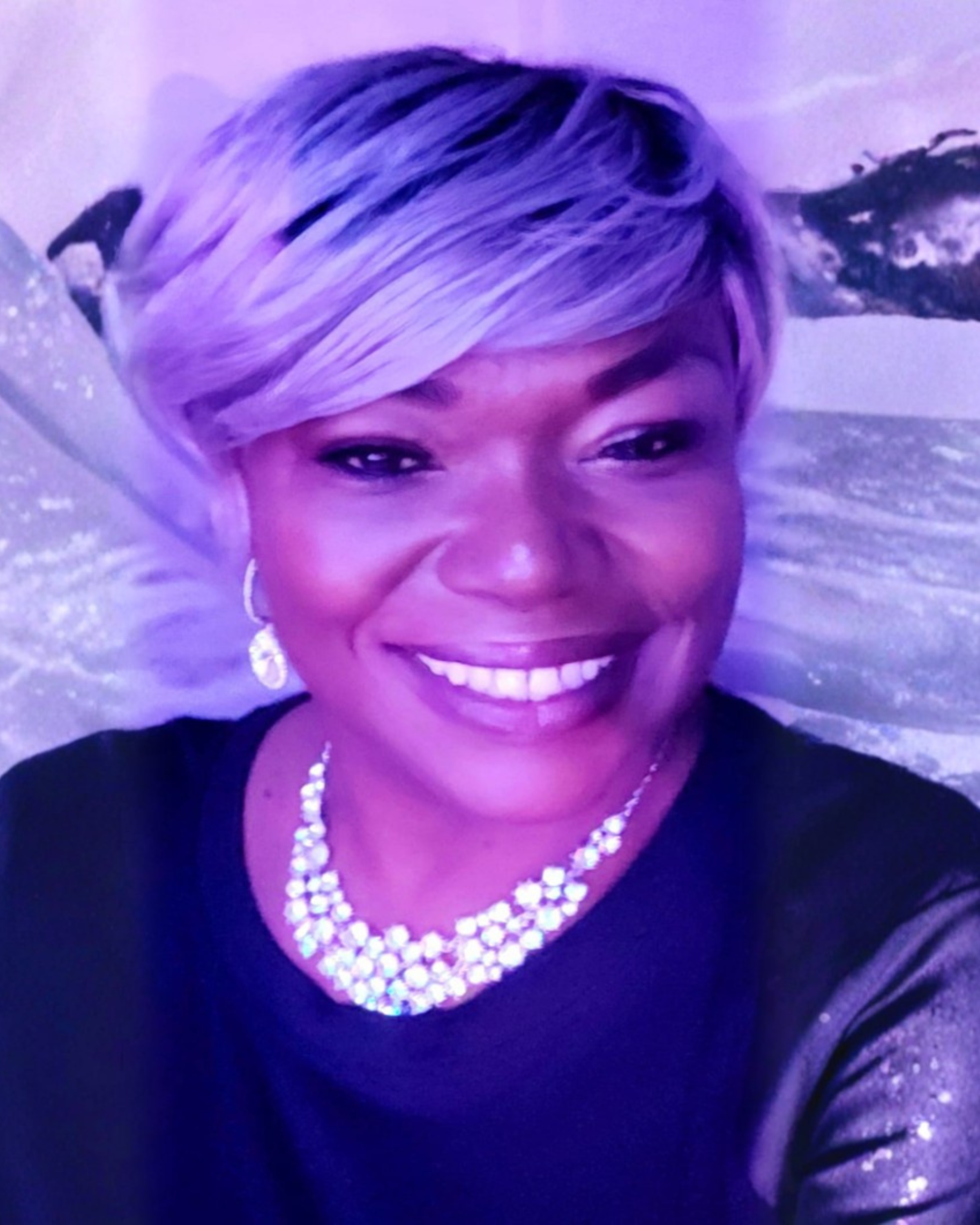Smiling woman with short blonde hair wearing a pearl necklace and earrings, dressed in a black top, at a formal event with a purple-tinted background.
