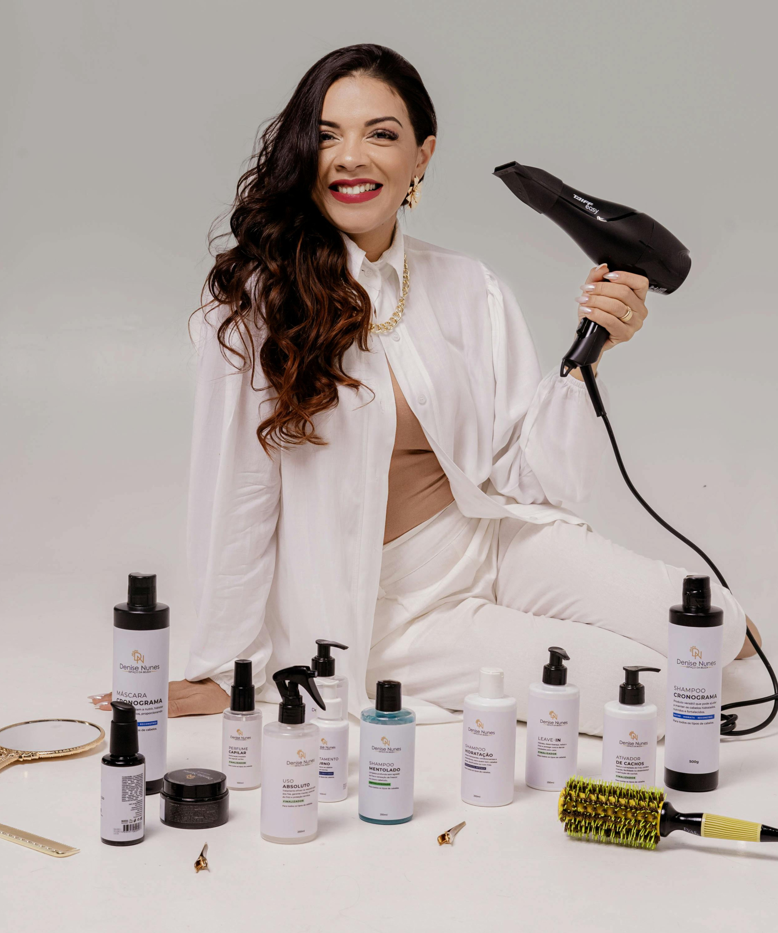A woman with long dark curly hair, wearing a white shirt and jewelry, smiling and holding a black hair dryer. In front of her are various hair care and styling products on a white surface.