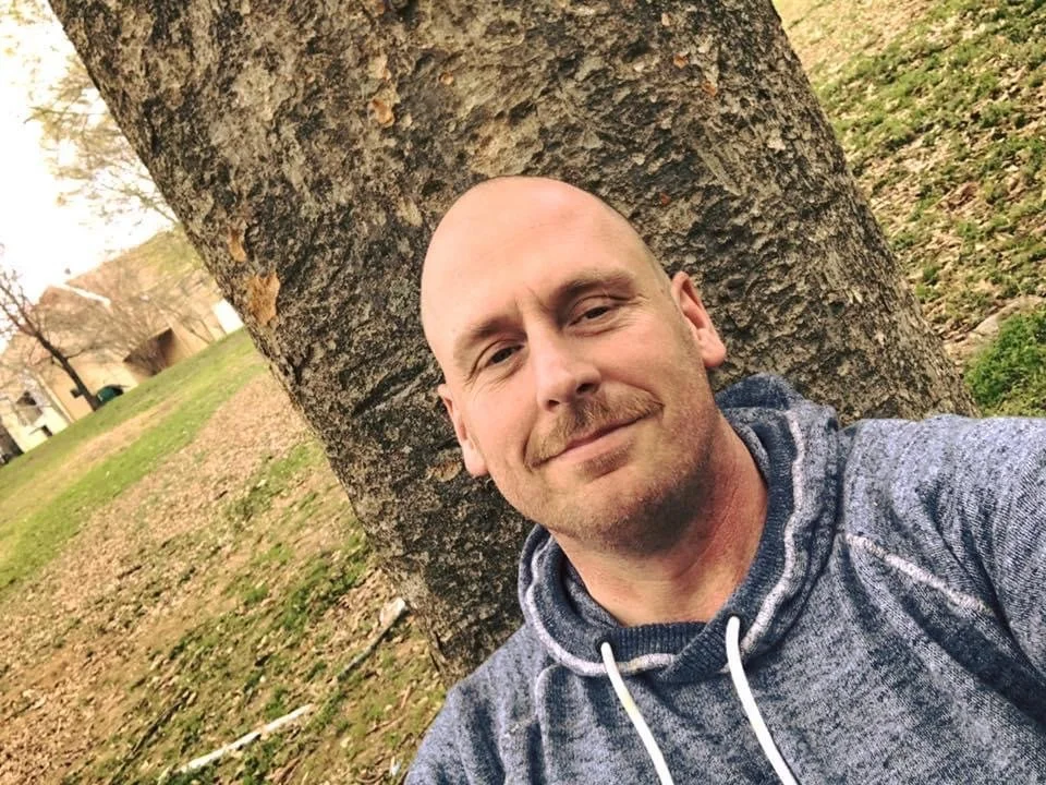 A man with a shaved head and goatee smiling while sitting next to a large tree in a park, wearing a gray hoodie.