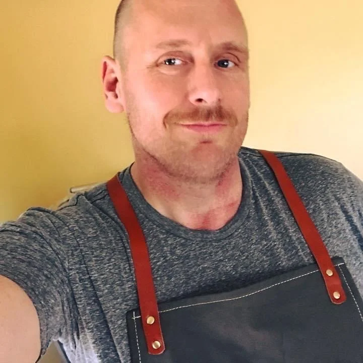 A man taking a selfie, wearing a grey t-shirt and a black apron with red straps, standing against a yellow wall.