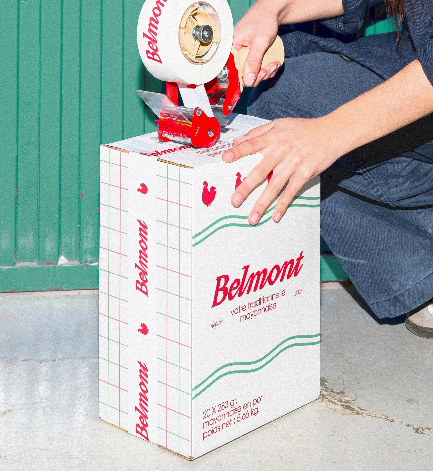 Personne scotchant un carton d'expédition Belmont avec illustrations rouges et vertes, posé sur sol béton devant mur vert — packaging produit alimentaire par Deux lignes Studio