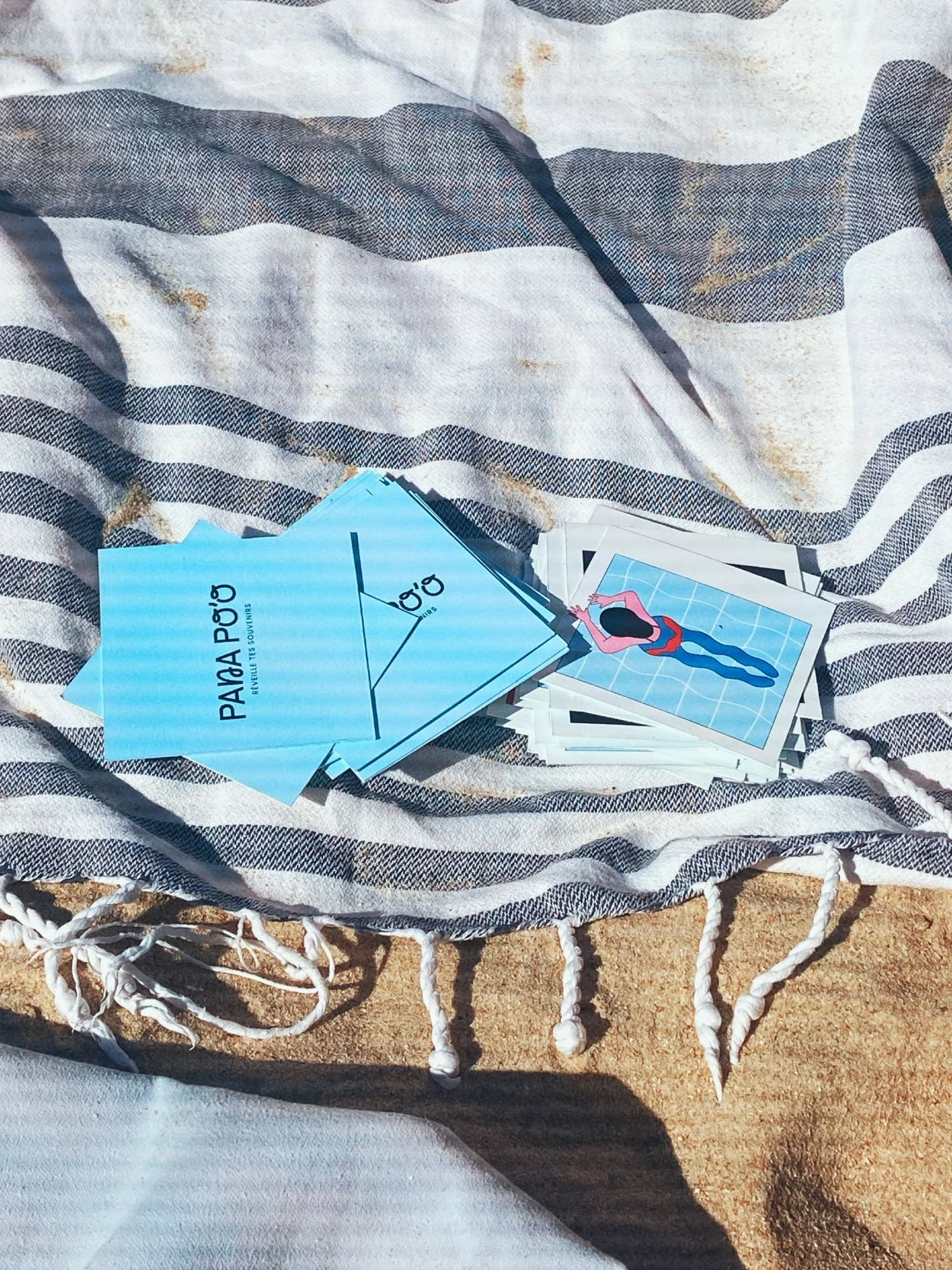 Pile de cartes colorées Pana po'o posées sur une serviette de plage rayée sur le sable — shooting lifestyle pour le jeu de cartes par Deux lignes Studio