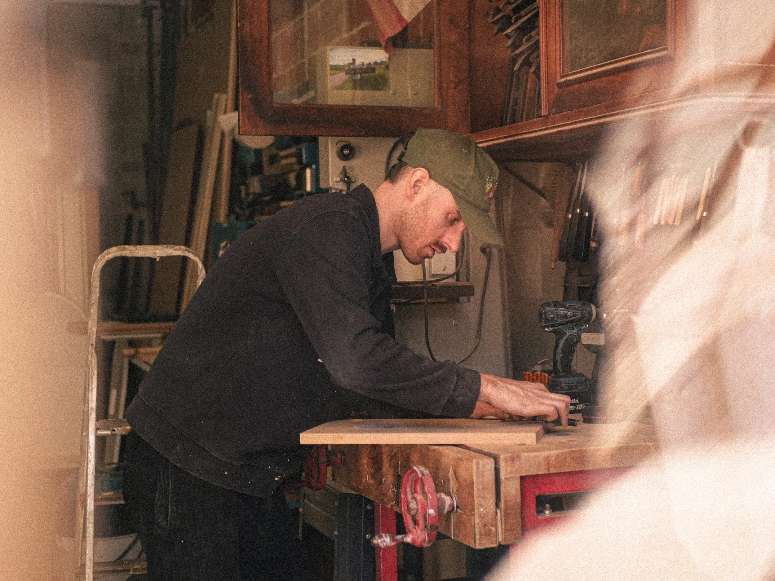 A man wearing a green cap and black shirt working in a woodworking shop, surrounded by tools and wood pieces.
