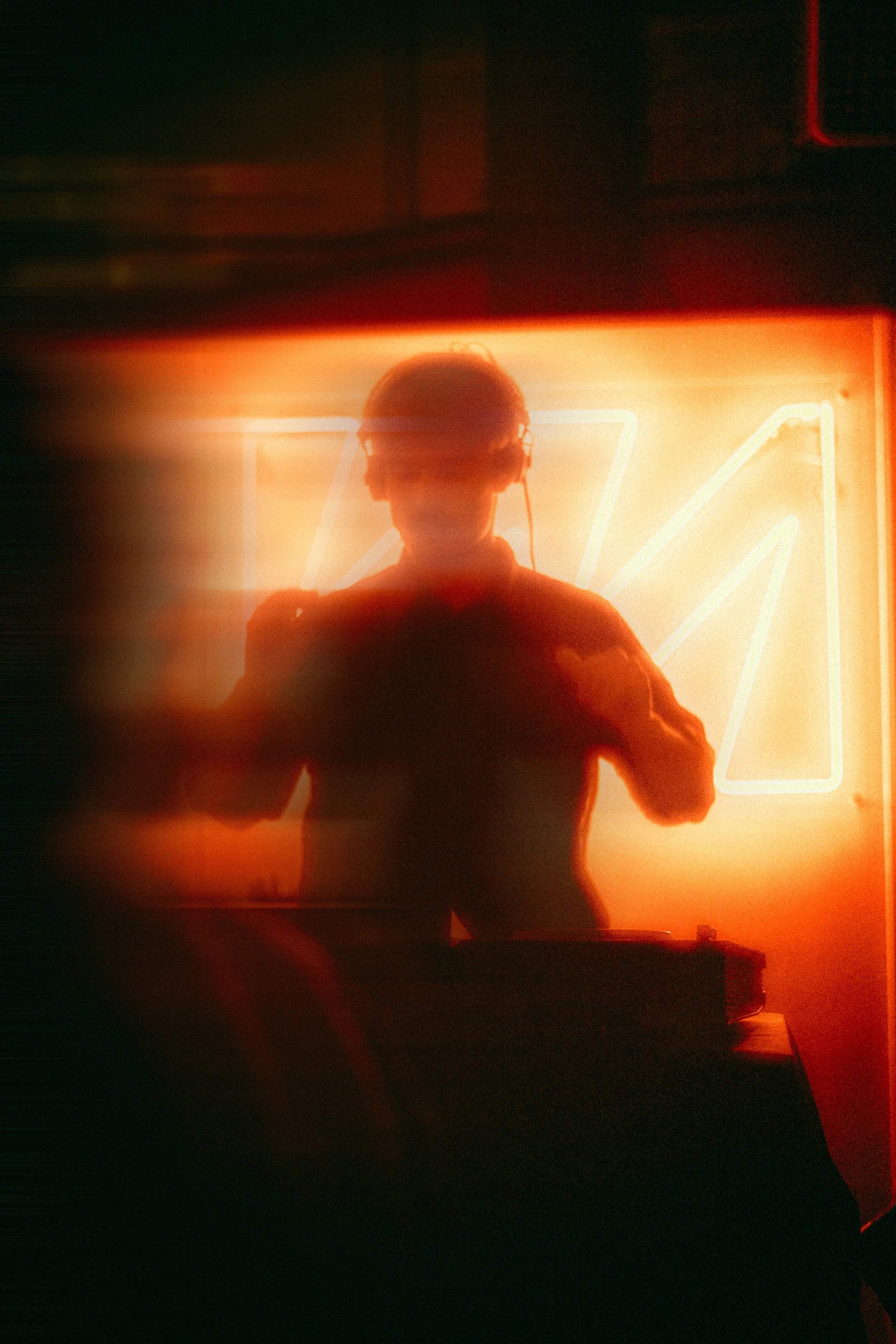 A person with headphones standing in front of a neon light with a geometric design, in a dark setting.