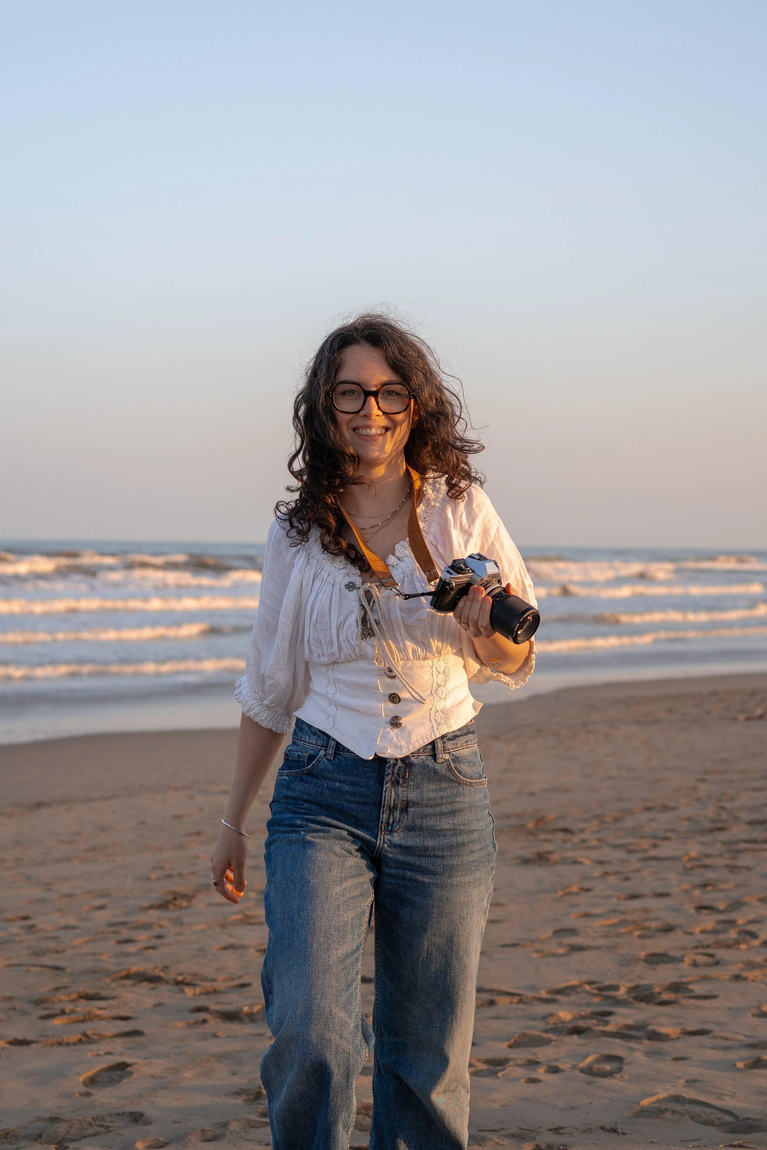 A woman with curly hair, glasses, and a white blouse holding a camera, standing on a sandy beach during sunset, with waves in the background.