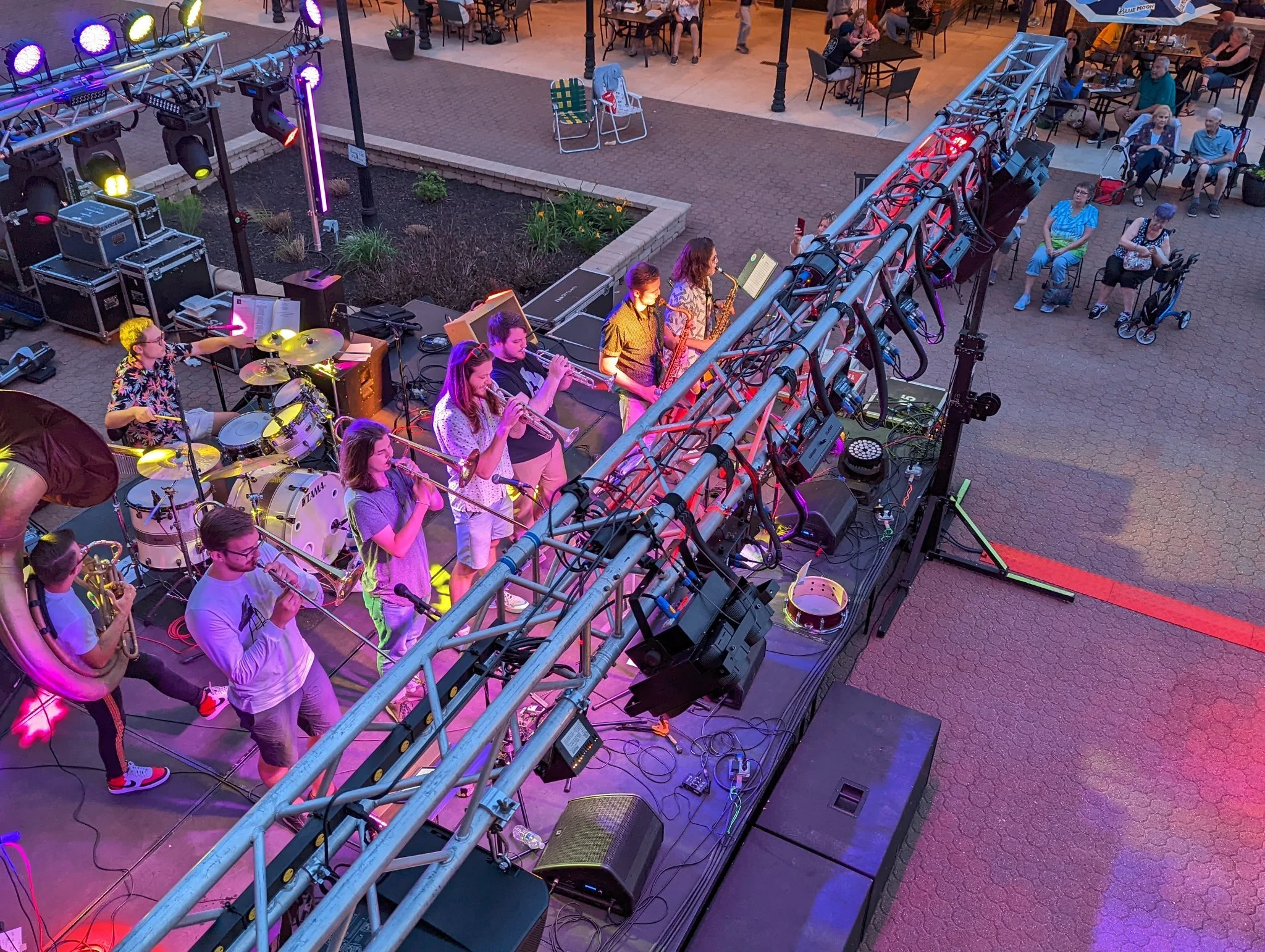 An overhead view of a live outdoor band performance at dusk, with musicians playing instruments such as drums, saxophones, and trumpet on a stage with purple and yellow lighting. Audience members are seated at tables and some are taking photos.