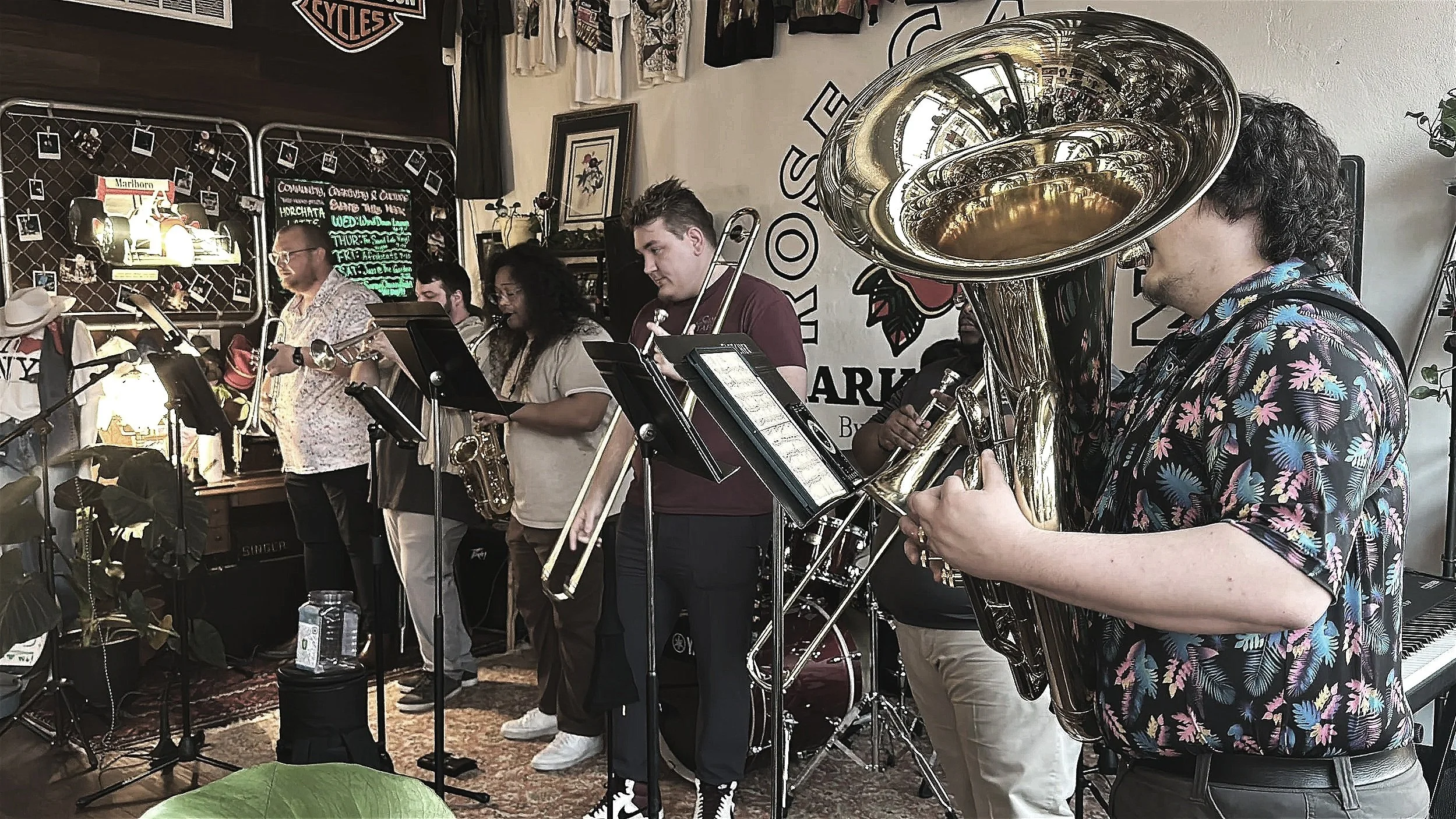 A group of musicians playing instruments, including a saxophone, trombone, and tuba, during a live performance in a cozy venue decorated with posters, photographs, and artwork on the walls.