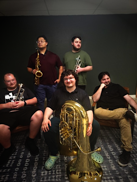 Five young men with musical instruments in an indoor setting with a dark wall background. One seated holding a baritone saxophone, two standing holding alto saxophones, and two seated on either side, all smiling or relaxed.