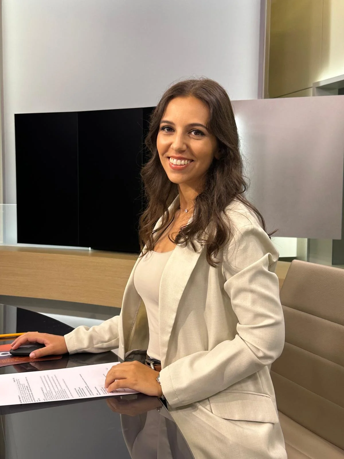 Mulher sentada em uma mesa de escritório, sorrindo, com documentos na mão e usando blazer bege.
