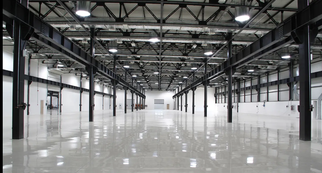 Empty industrial warehouse with bright lighting and high ceilings, featuring black steel support beams and polished concrete floors.