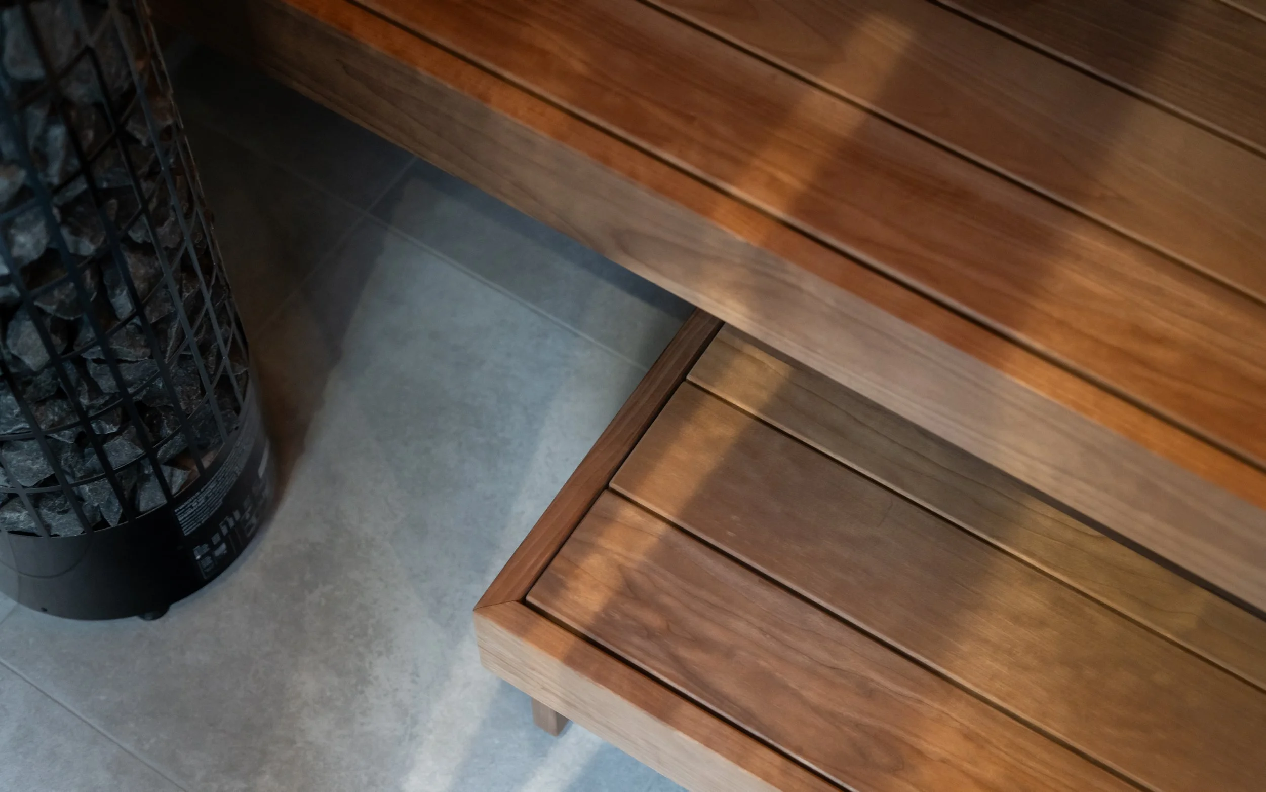 A wooden bench with horizontal slats, placed in a room with tiled flooring. A cylindrical container filled with rocks or similar material is positioned next to the bench.