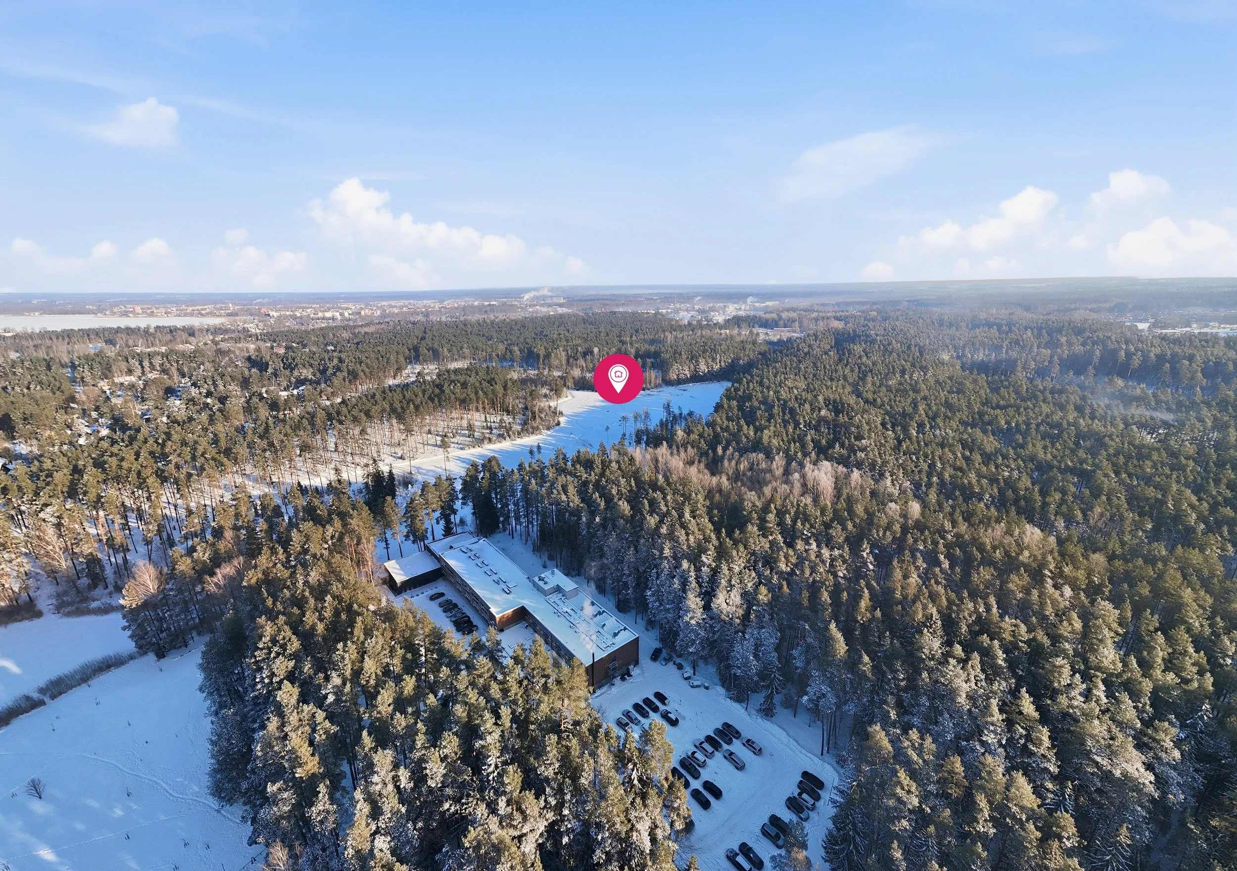 A snow-covered forest with a building and parking lot at the bottom, and a distant cityscape with a blue sky and clouds.