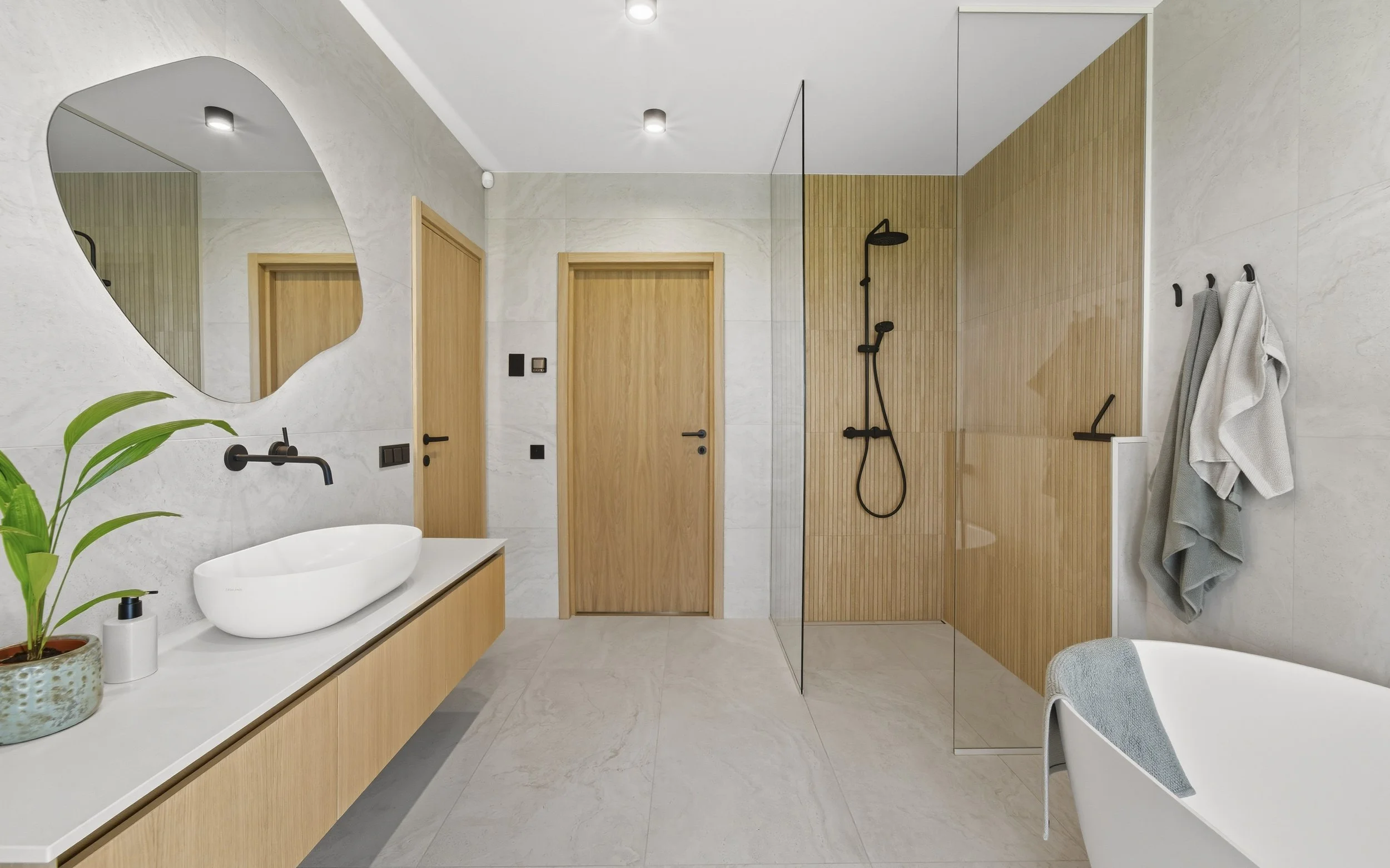 Modern bathroom with a white sink on a wooden vanity, black faucet, large mirror, potted plant, wooden doors, walk-in shower with black fixtures and a wood-paneled accent wall, towels on hooks, and a white chair with a towel.