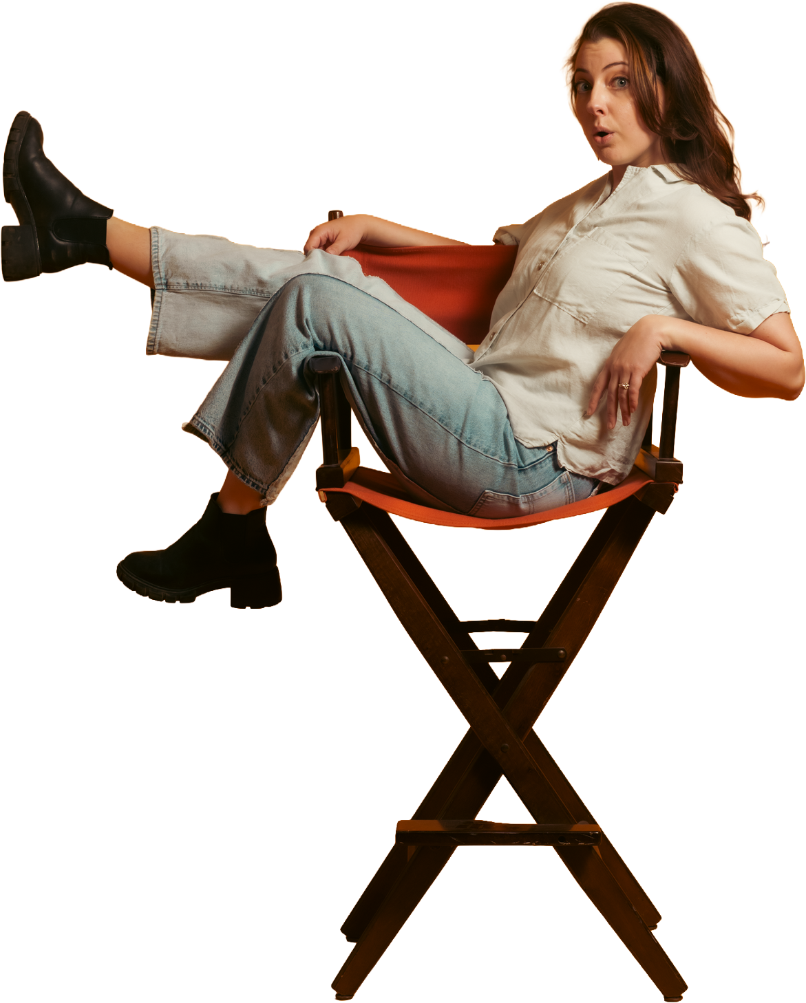 A woman seated on a director's chair with her leg stretched out, wearing black boots, light-washed jeans, a beige short-sleeve shirt, and with red hair. She is looking at the camera with a surprised expression.