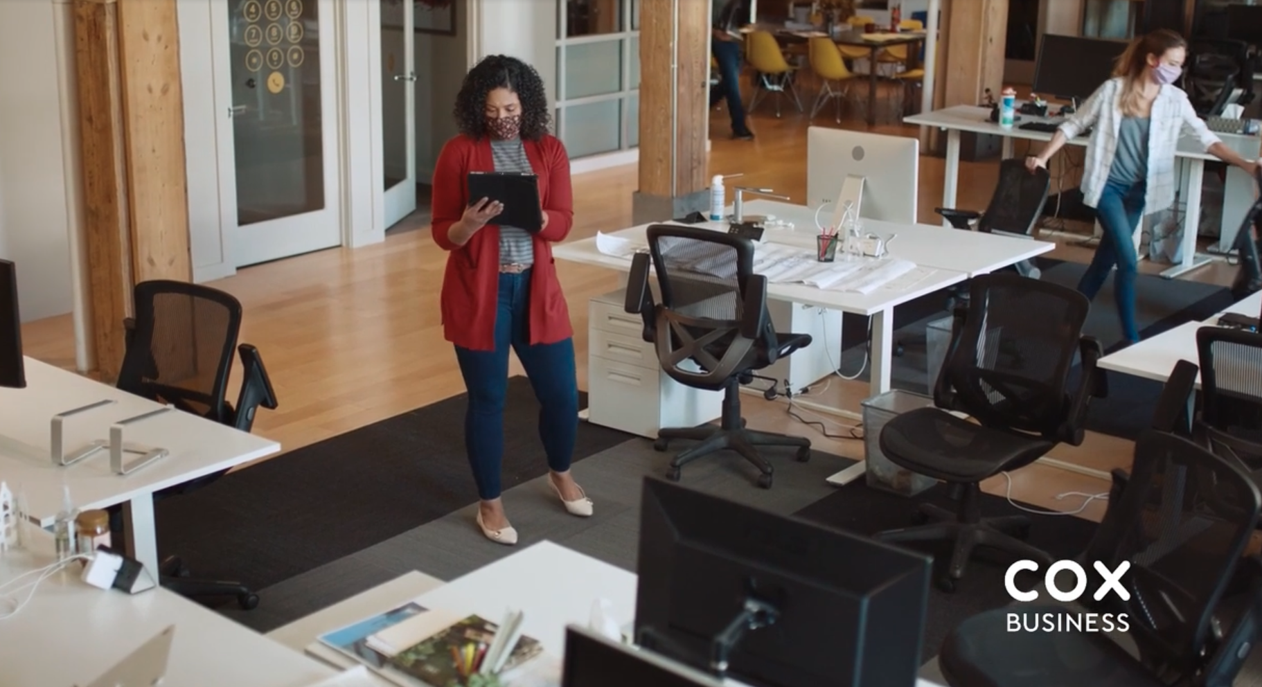 An office space with women wearing masks. One woman stands at the center holding a tablet, wearing a red blazer, striped shirt, blue jeans, and beige heels. Two other women are seen in the background, one walking and one near a desk.