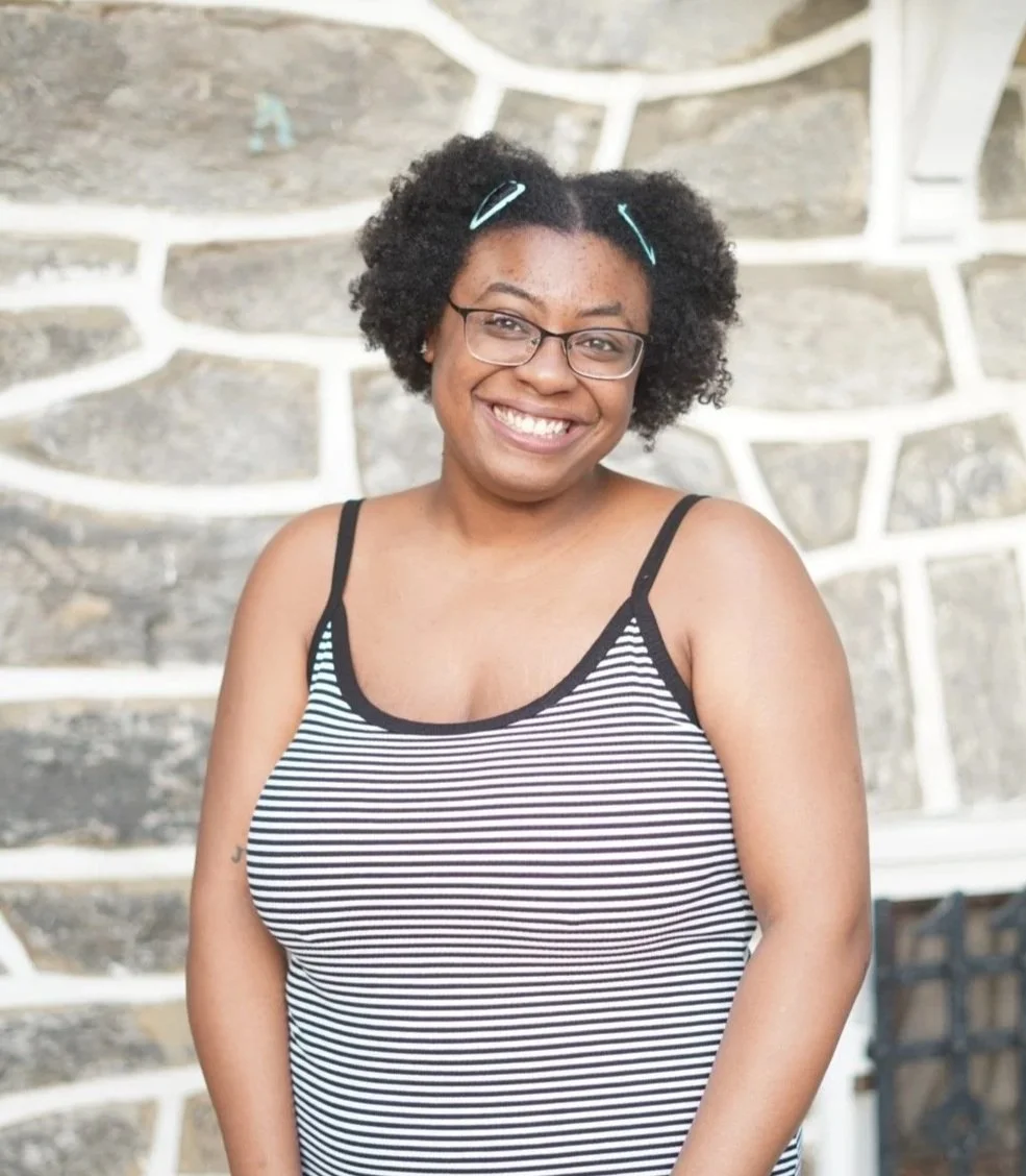 Smiling woman with glasses and curly hair, wearing a striped tank top, standing outdoors in front of a stone wall.