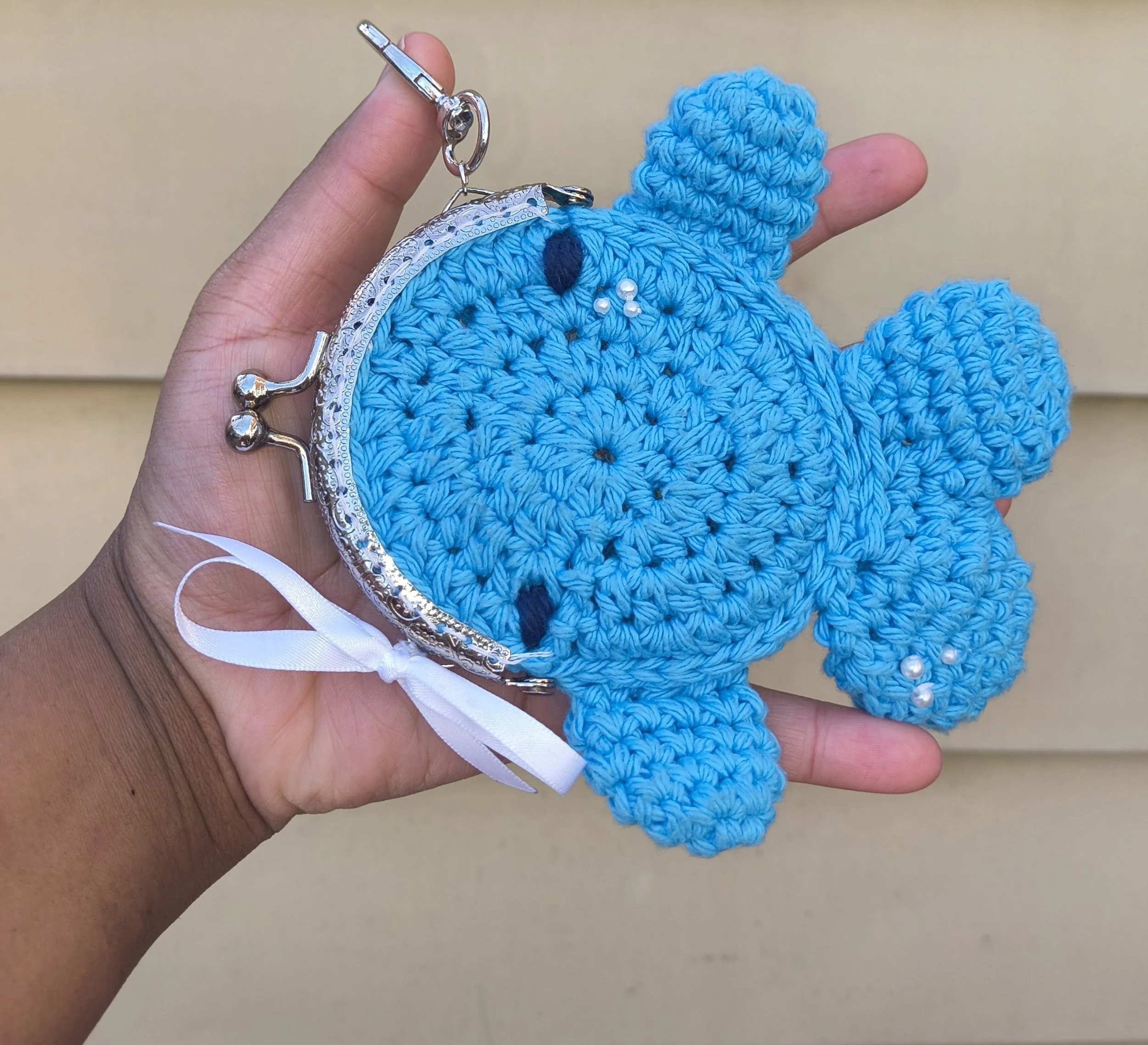 A hand holding a blue crocheted elephant coin purse with a silver frame and a white ribbon tied on the side.