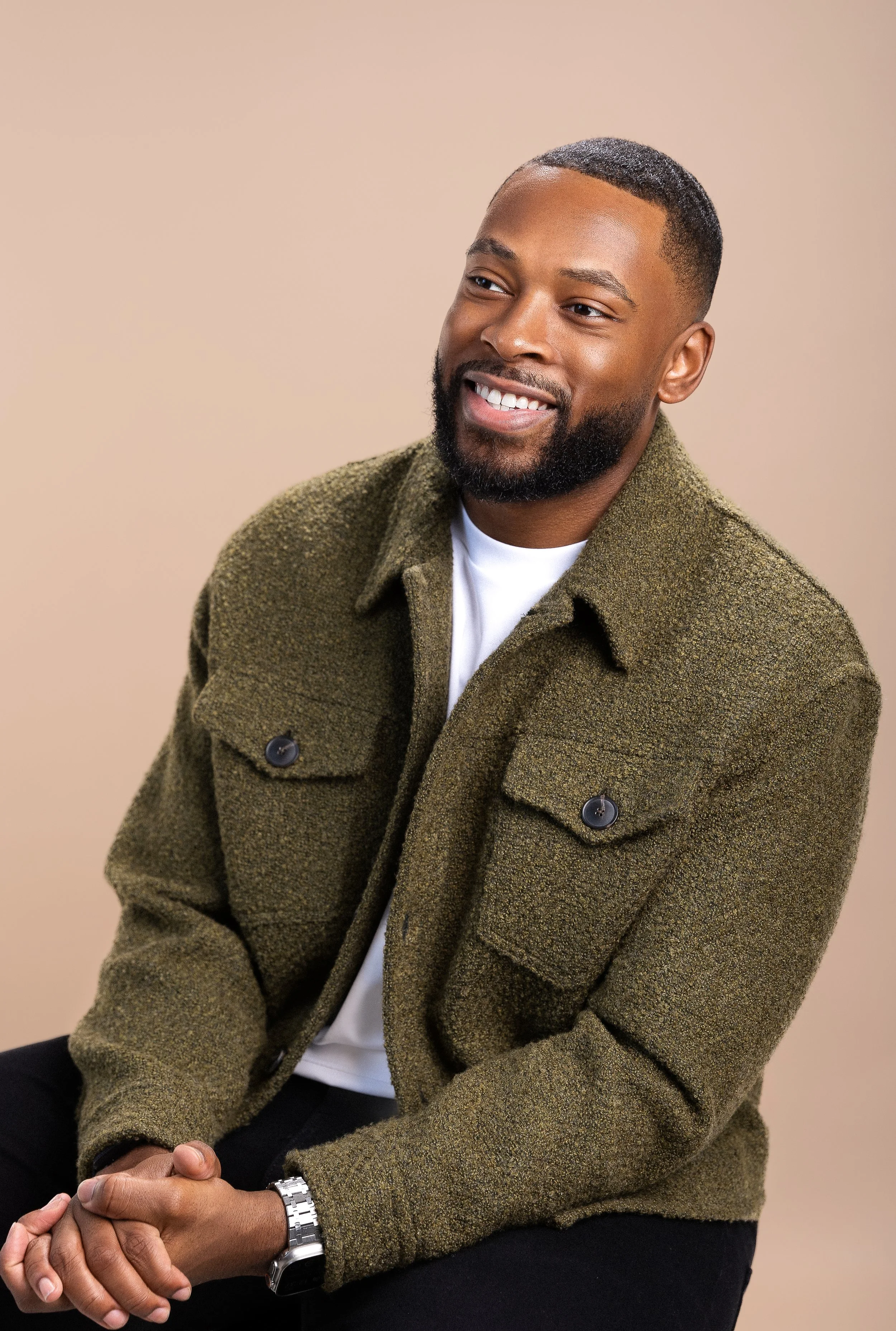 Brandon Smithwrick smiling and wearing a greenish-brown coat over a white shirt. He is sitting with his hands clasped in front of him and a silver watch on his left wrist, against a plain beige background.