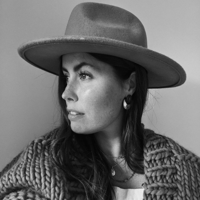 Adrienne Sharpe wearing a wide-brimmed hat, earrings, layered necklaces, and a chunky knit sweater, looking to the side in black and white.
