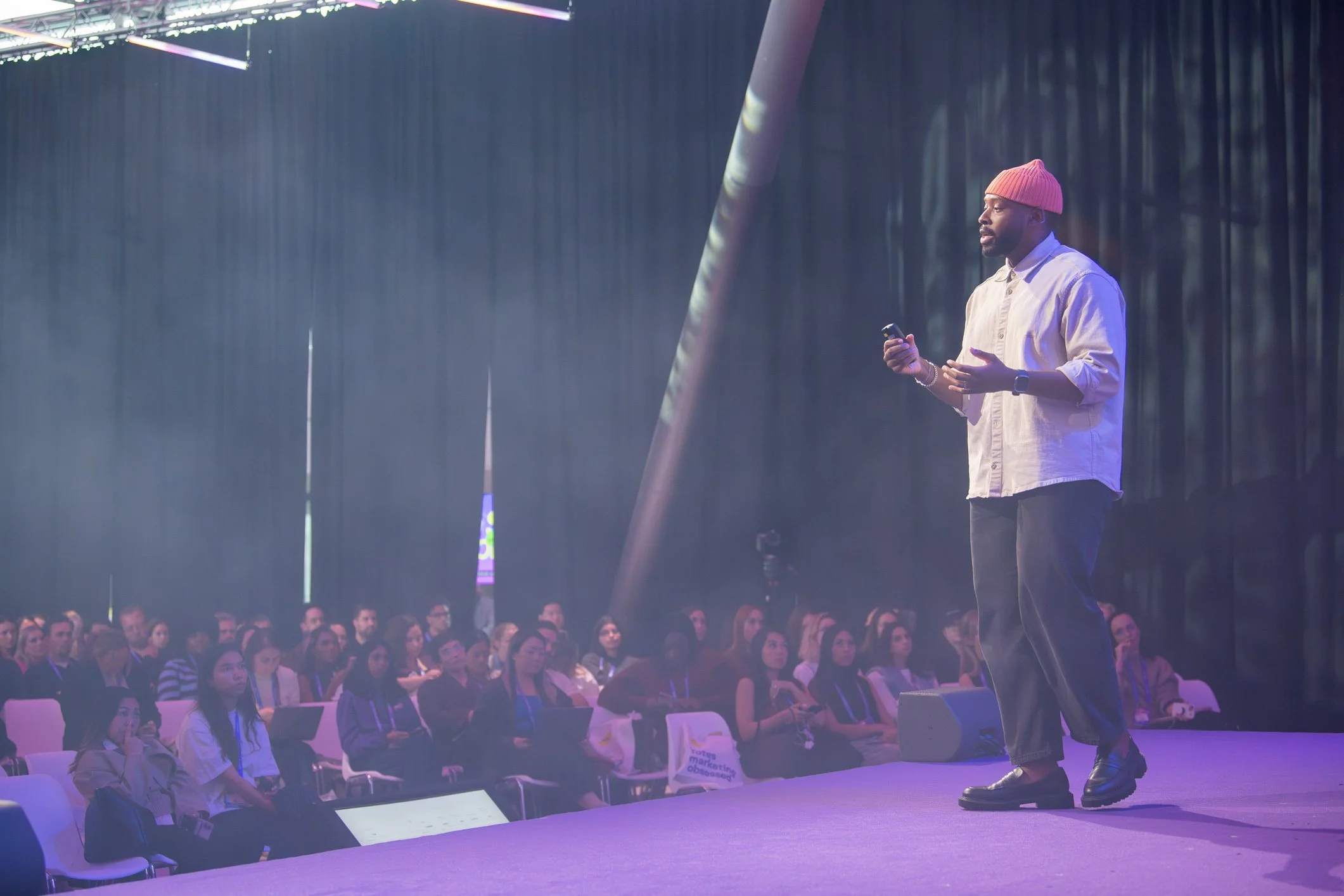 Brandon Smithwrick in an orange beanie, and beige shirt giving a presentation to an audience in a large dimly lit conference room with colored stage lights.