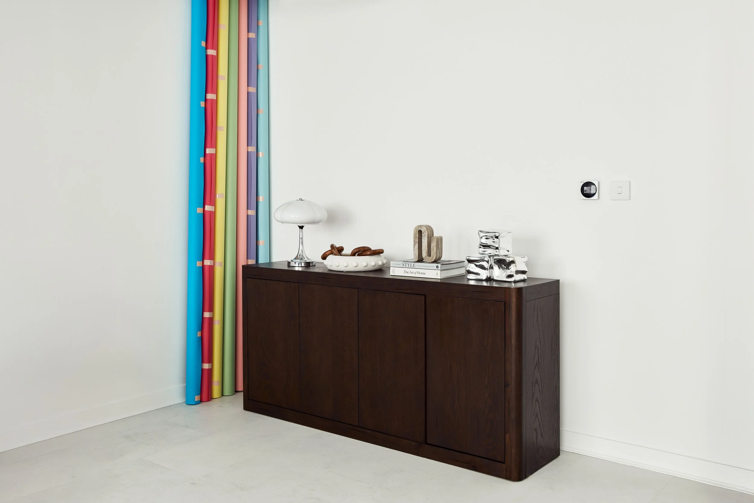 Dark wooden sideboard with decorative items, including a white lamp, books, and abstract sculptures, beside a colorful coloramas against a plain white wall.