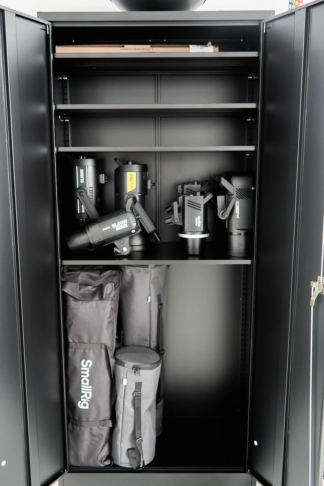Black storage locker containing lighting equipment, including four black professional stage or studio lights on the middle shelf and two large black bags on the bottom shelf.