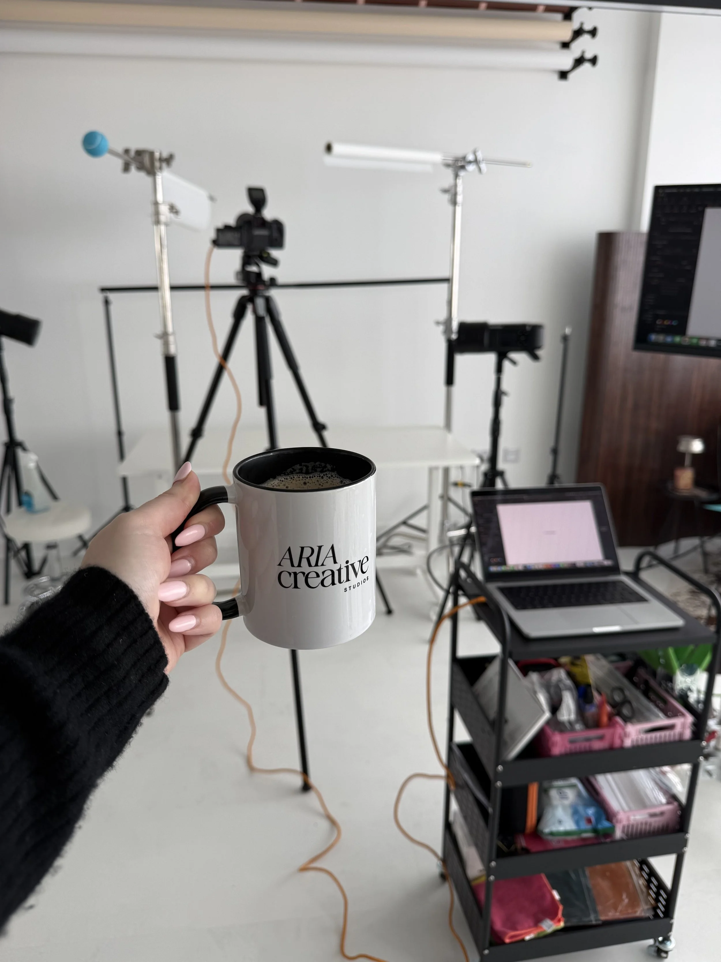 A person holding a white coffee mug with black text that says 'ARIA creative Studios' in a photography or filming studio, with lighting equipment, cameras, a laptop, and a rolling cart with supplies in the background.