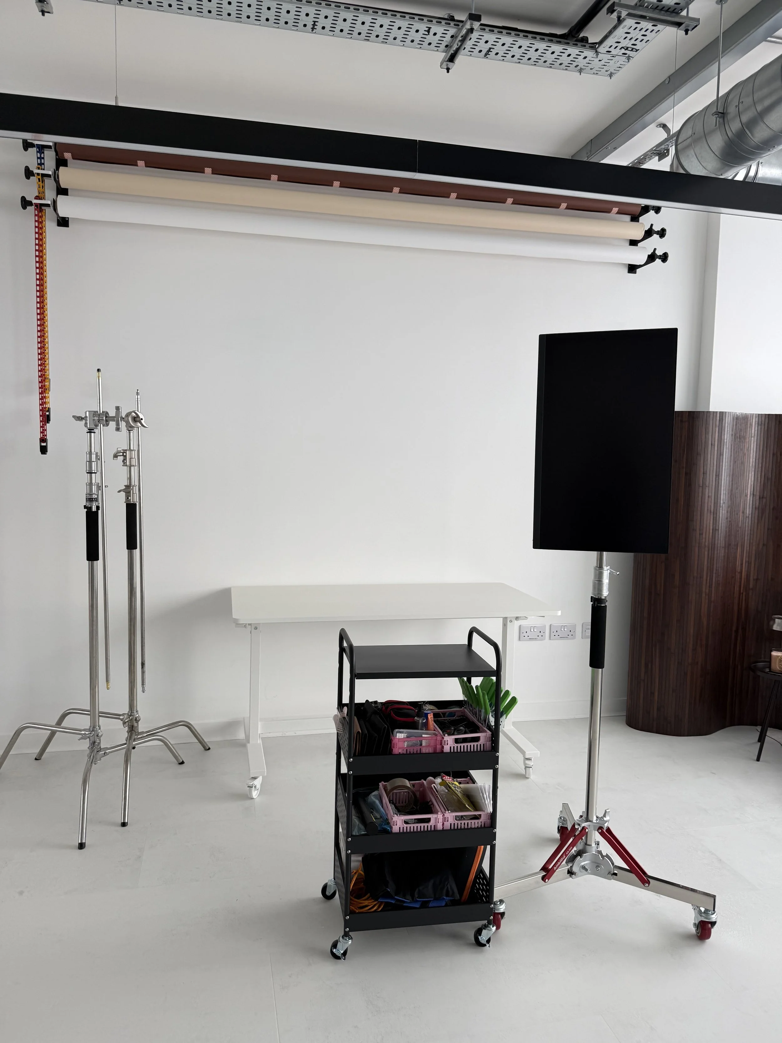 Photo of a photography studio setup with backdrop rolls, lighting equipment, a rolling cart with tools, and a large screen on a stand.