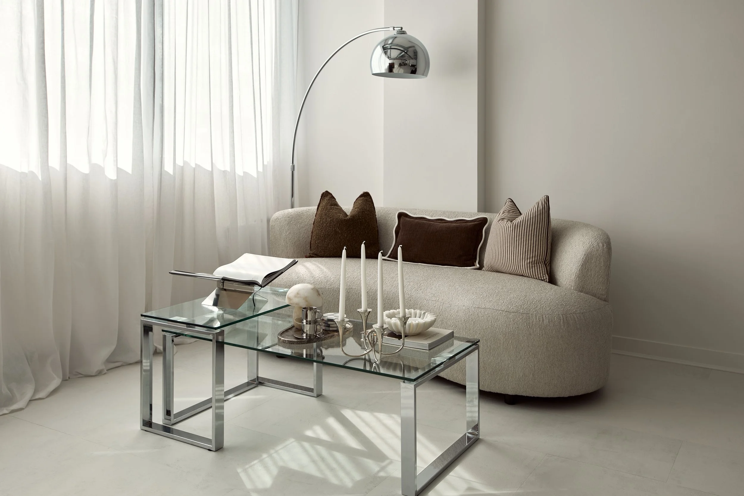 Minimalist living room with a beige sofa, brown and striped accent pillows, a glass coffee table with a book and decorative items, and a chrome arc floor lamp, all in a bright, white space with curtains.