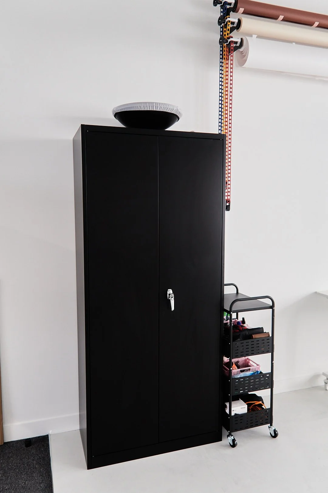 Black cabinet with a locked door, on top of which is a black and white bowl. To the right of the cabinet, a three-tier rolling cart with various items. Above, some colored measuring tapes and rolls of paper are mounted on the wall.
