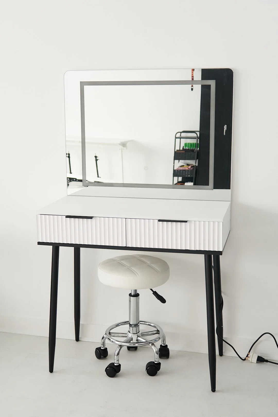White vanity table with a large mirror and a padded stool in front, set against a white wall.