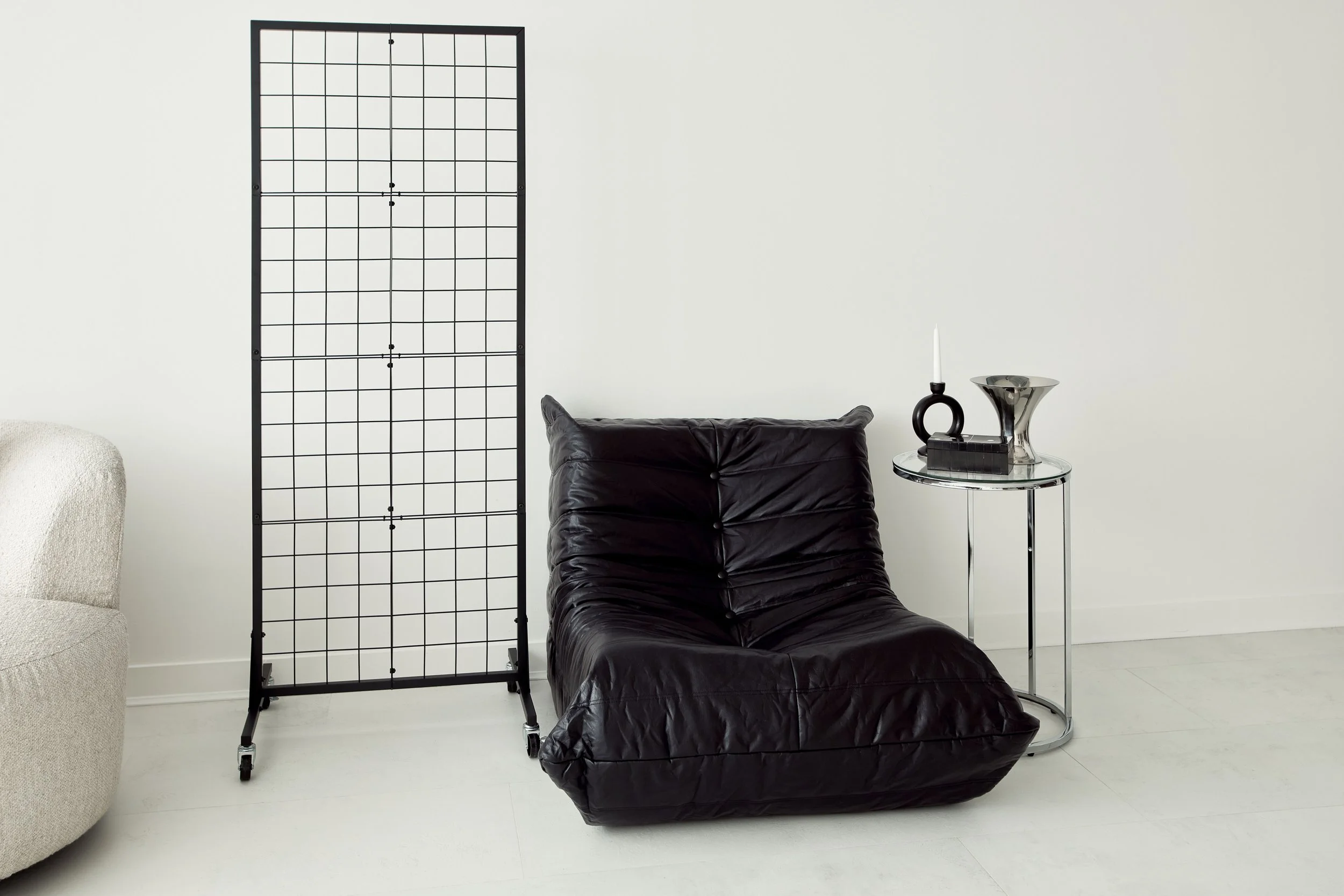 Minimalist living room corner with a beige armchair, a black leather lounge chair, a glass side table with a black candle holder, a candle, a decorative vase, and a grid room divider, all against a white wall.