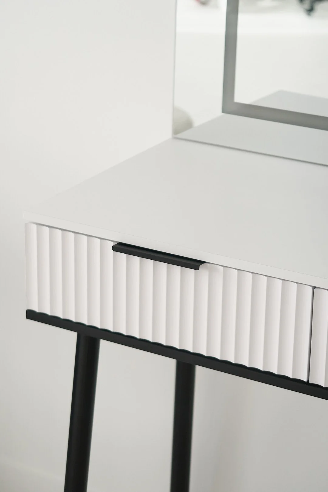Close-up of a white modern side table with vertical ridges on the drawer front, black handle and black legs, with a mirror reflecting part of a white wall in the background.