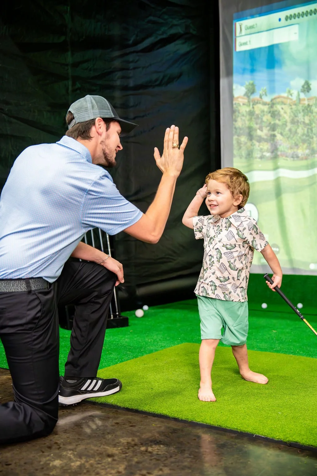 A man is giving a high five to a young boy who is holding a golf club playing on Par-Tee Time's mobile golf simulator.