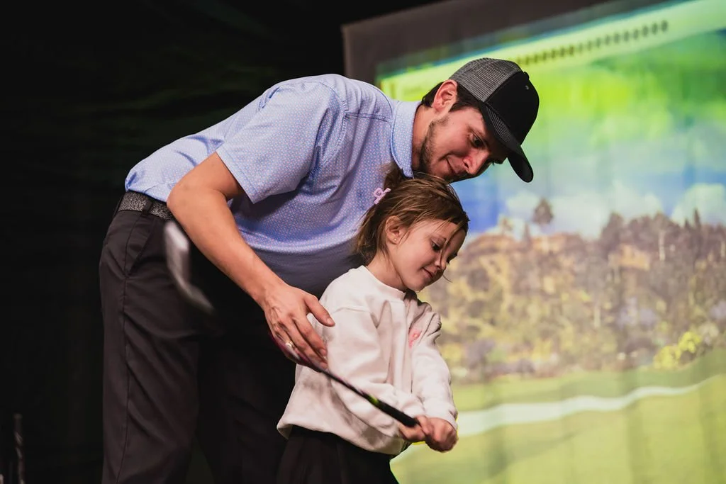 Dad teaching a young girl how to golf on a golf simulator with a large screen displaying a golf course in the background.