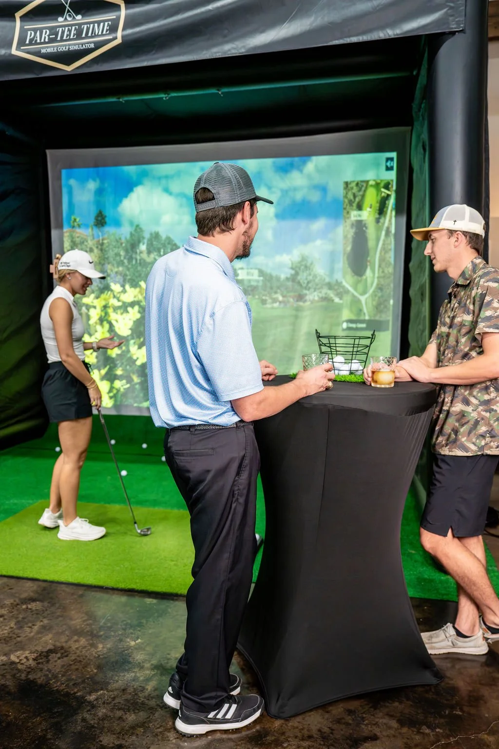 Three people standing at a high-top table with drinks, watching a golf simulation game projected on a screen, while a woman practices golf swings in a simulator room with a green turf area and screen background of a golf course.