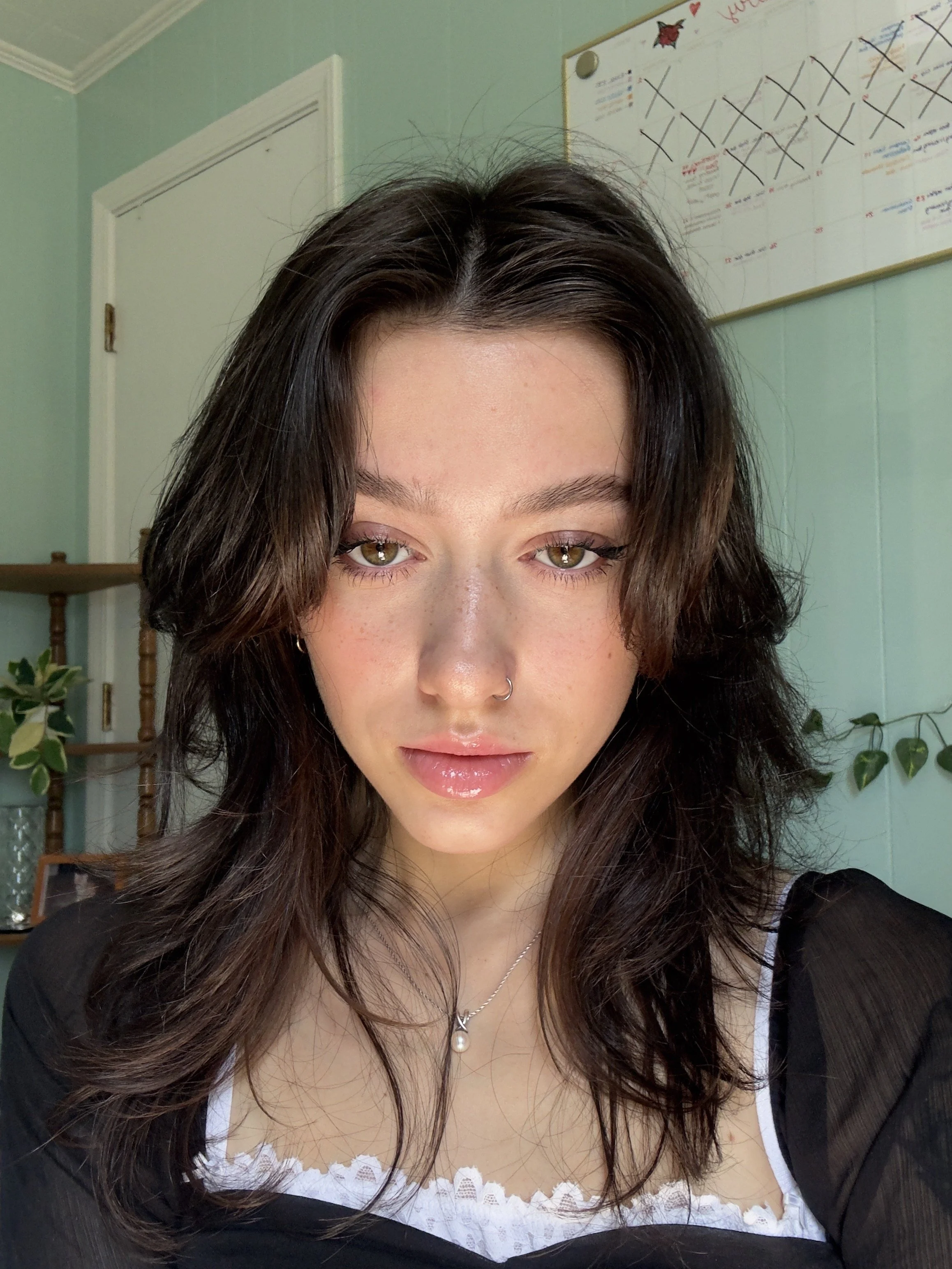 A woman with wavy brown hair, light skin, and hazel eyes, wearing a black top with lace trim, a silver necklace with a pearl pendant, in a room with green walls, plants, and a whiteboard in the background.