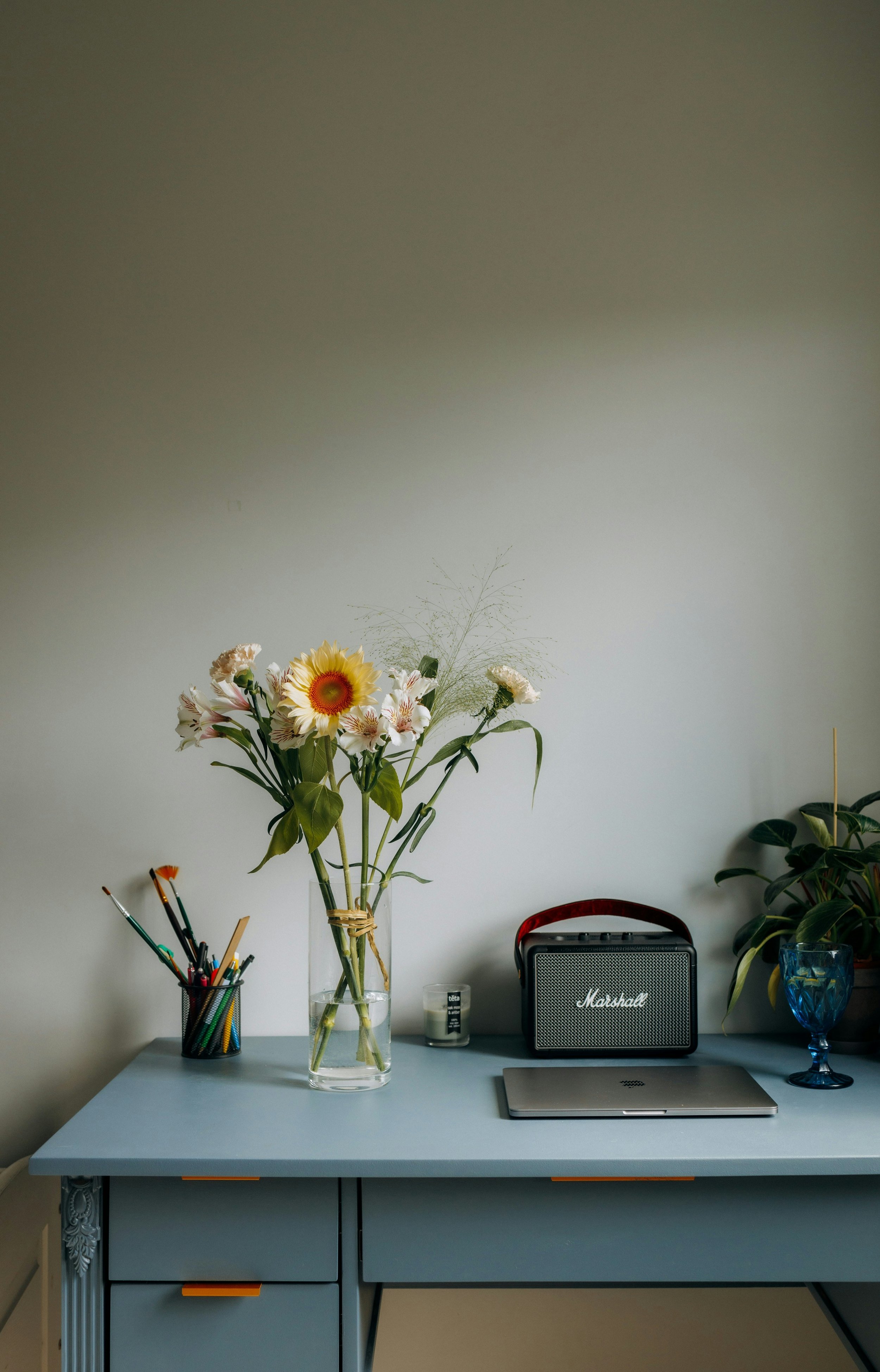 A blue desk with a clear glass vase holding a bouquet of flowers including sunflowers and lilies, a black pencil holder with colorful pens and brushes, a small candle, a Marshall portable speaker, a closed laptop, and a potted plant with large green leaves in a blue glass container.
